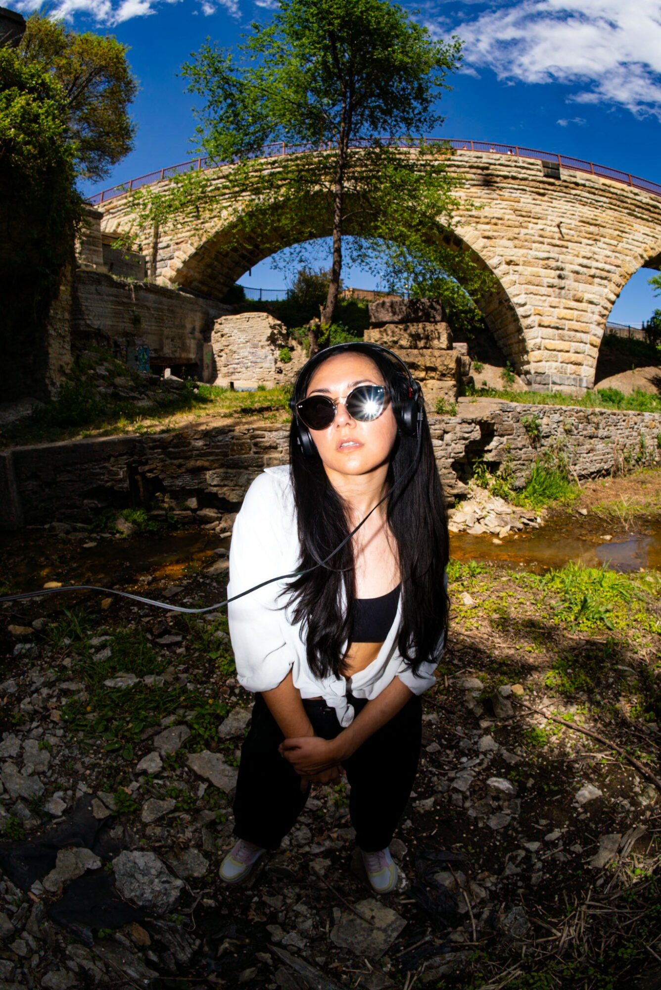 Young woman with long dark hair and sunglasses stands outdoors near an ancient stone bridge under a blue sky.