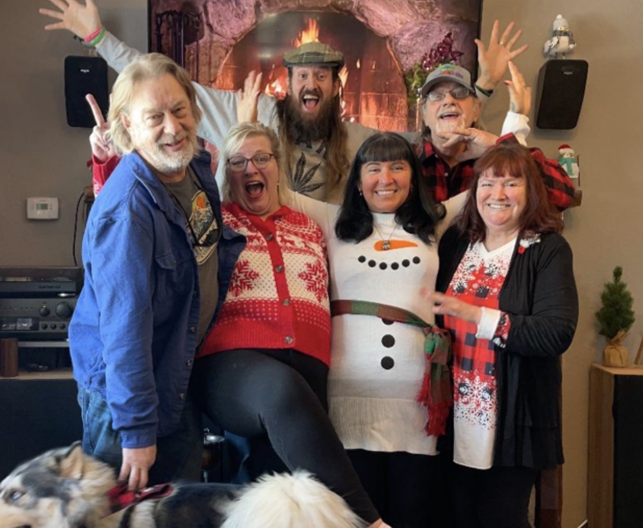 Group of six people celebrating in front of a fireplace, some wearing holiday sweaters, smiling and raising hands.
