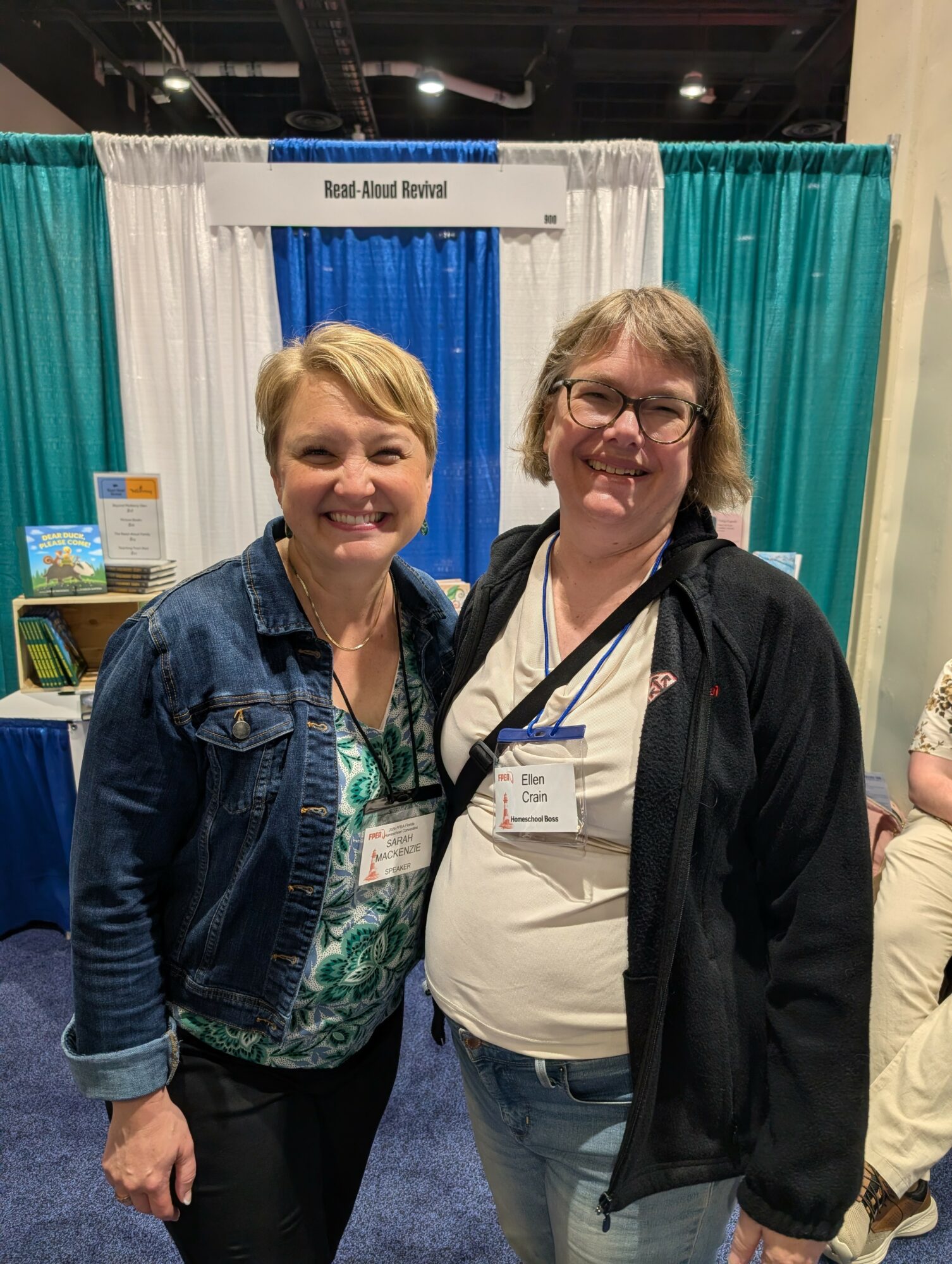 Two women smiling and standing together at an event booth with blue and teal curtains in the background.