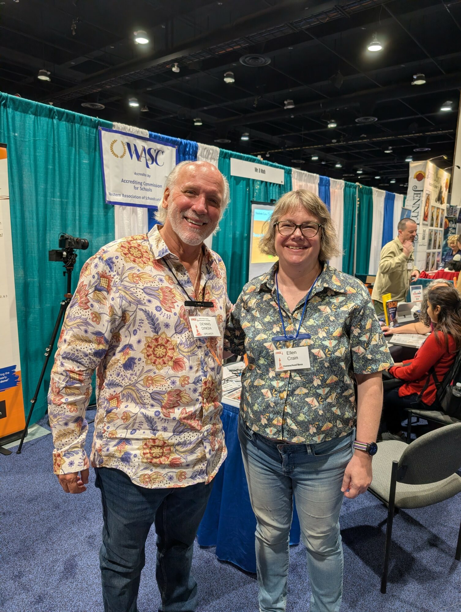 Two smiling people stand together at an indoor event with booths and banners in the background.