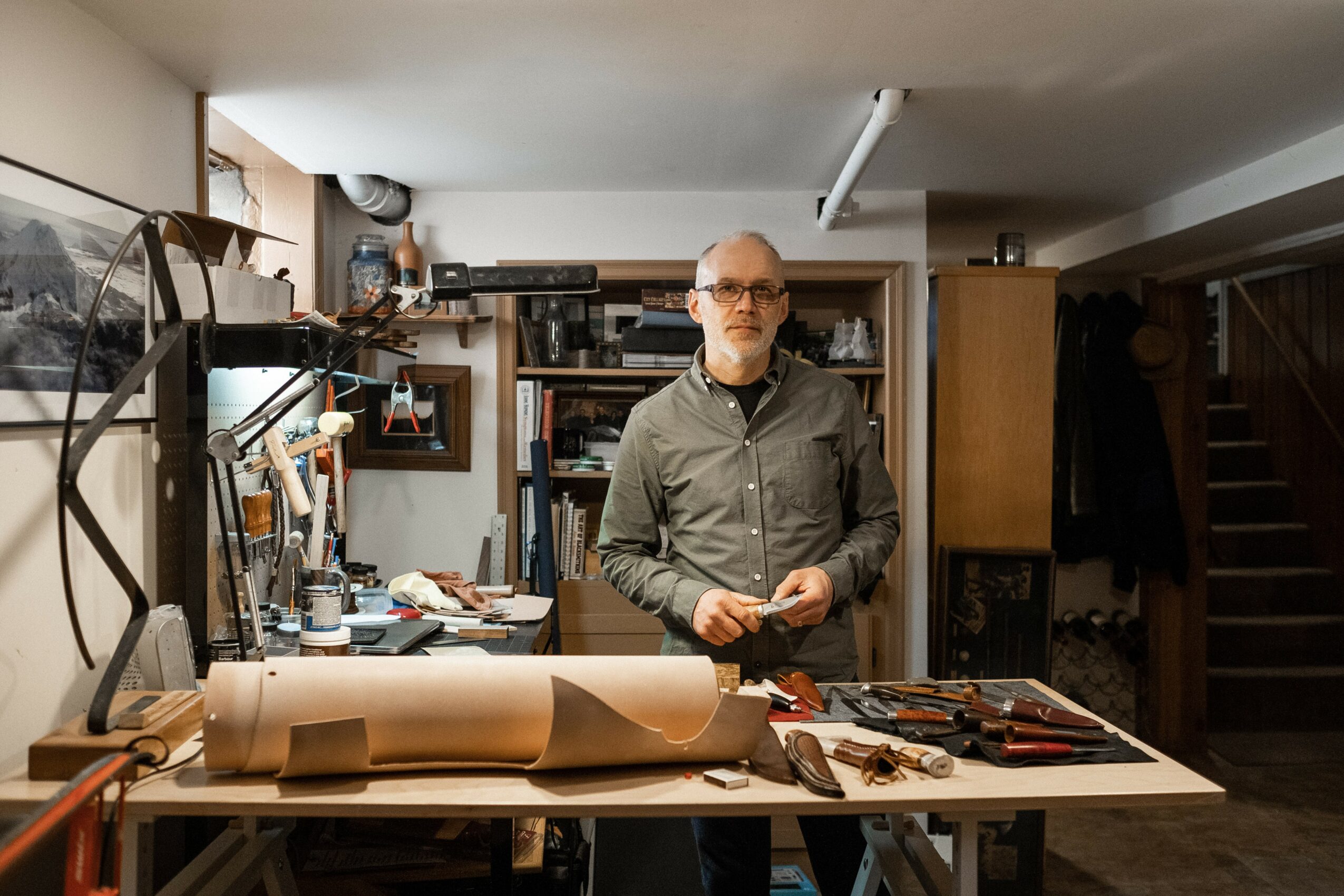 Man with glasses and gray shirt standing behind a worktable in a workshop, with tools and rolled paper on the table.