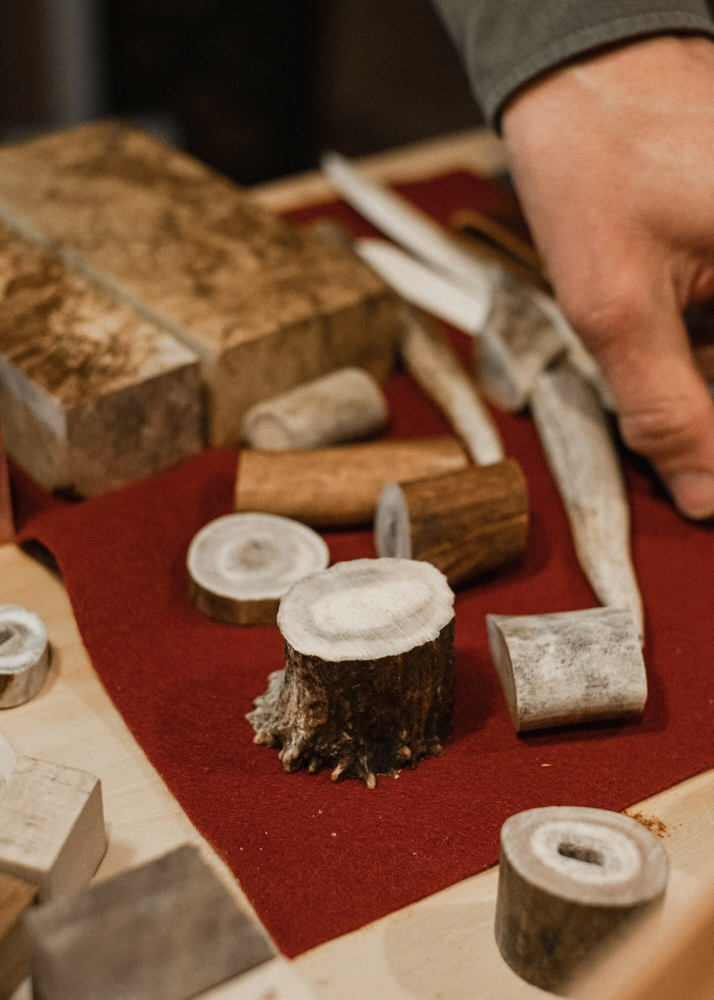 Person's hand near woodworking tools and wood pieces on red cloth, with a large block of wood in background.