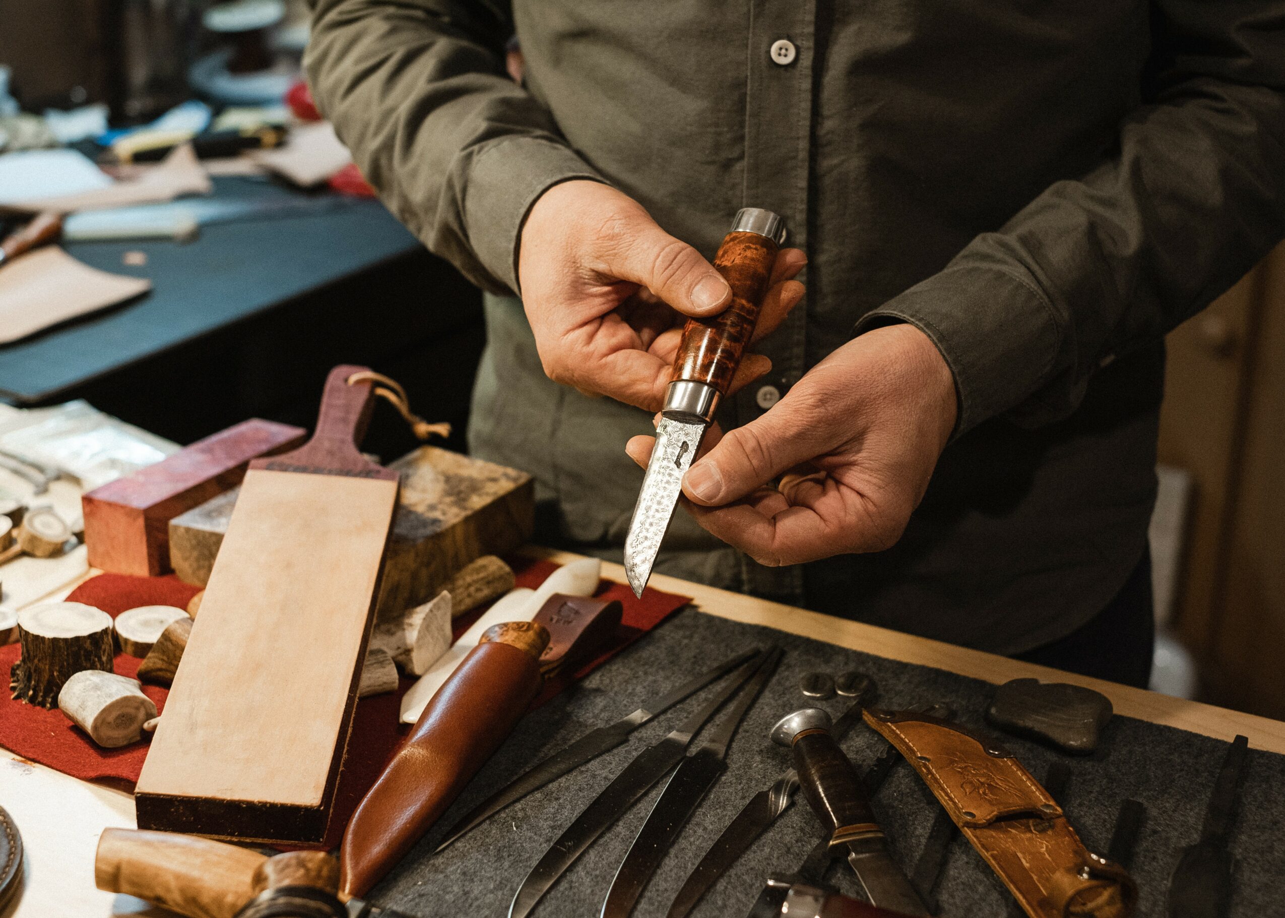 Person holding a carving knife and a wooden handle, surrounded by woodworking tools on a workbench.