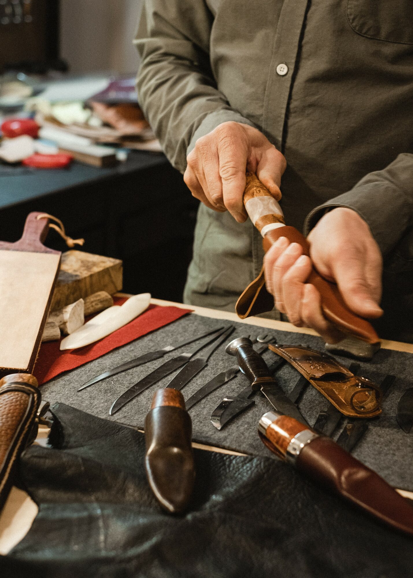 Person holding a striped knife handle over a workbench with various tools and leather items, workspace cluttered with supplies.