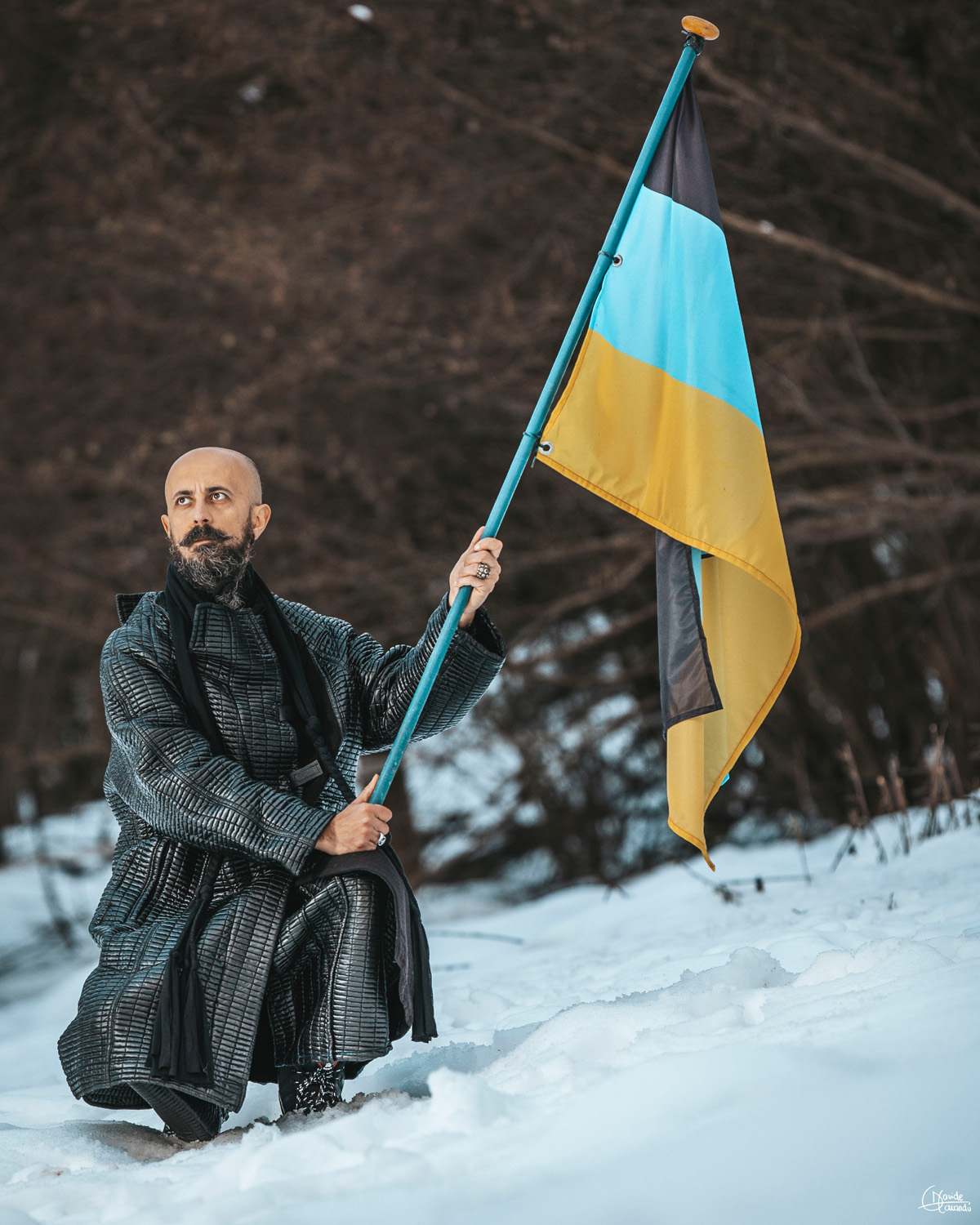Person kneeling in snow holding a Ukrainian flag outdoors, with trees in the background.