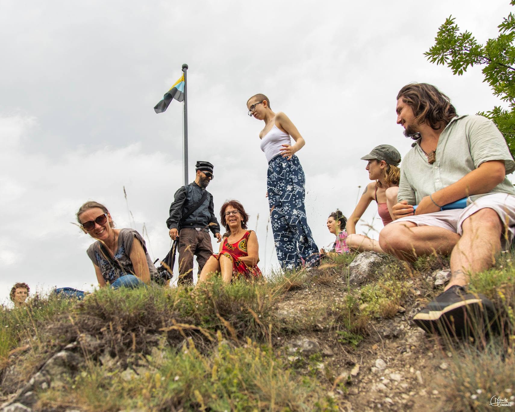 Group of people outdoors on a grassy hill with a flagpole, some sitting and others standing, under cloudy sky.