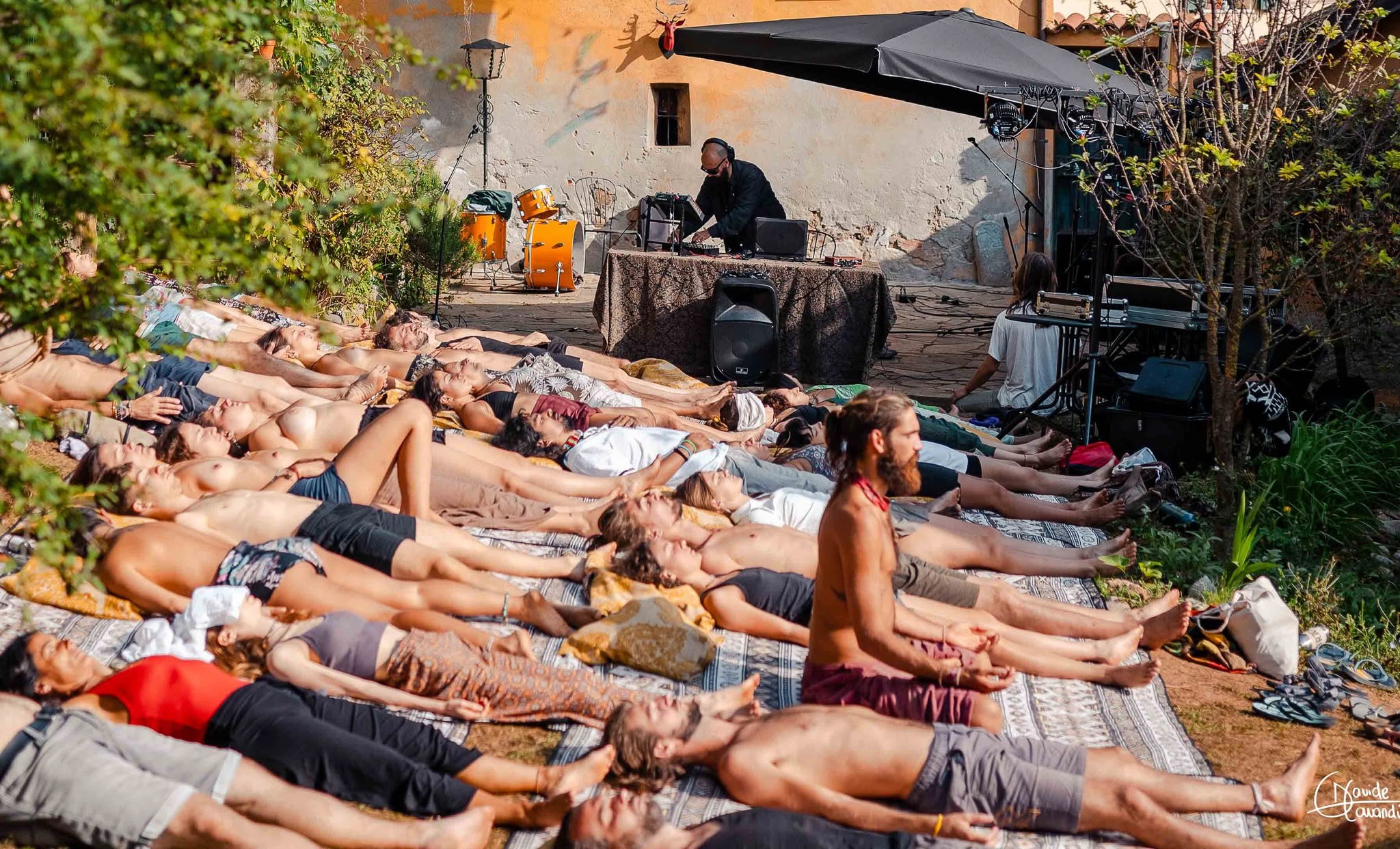 Group of people lying on mats outdoors, with a person sitting at a table in the background, surrounded by trees and plants.