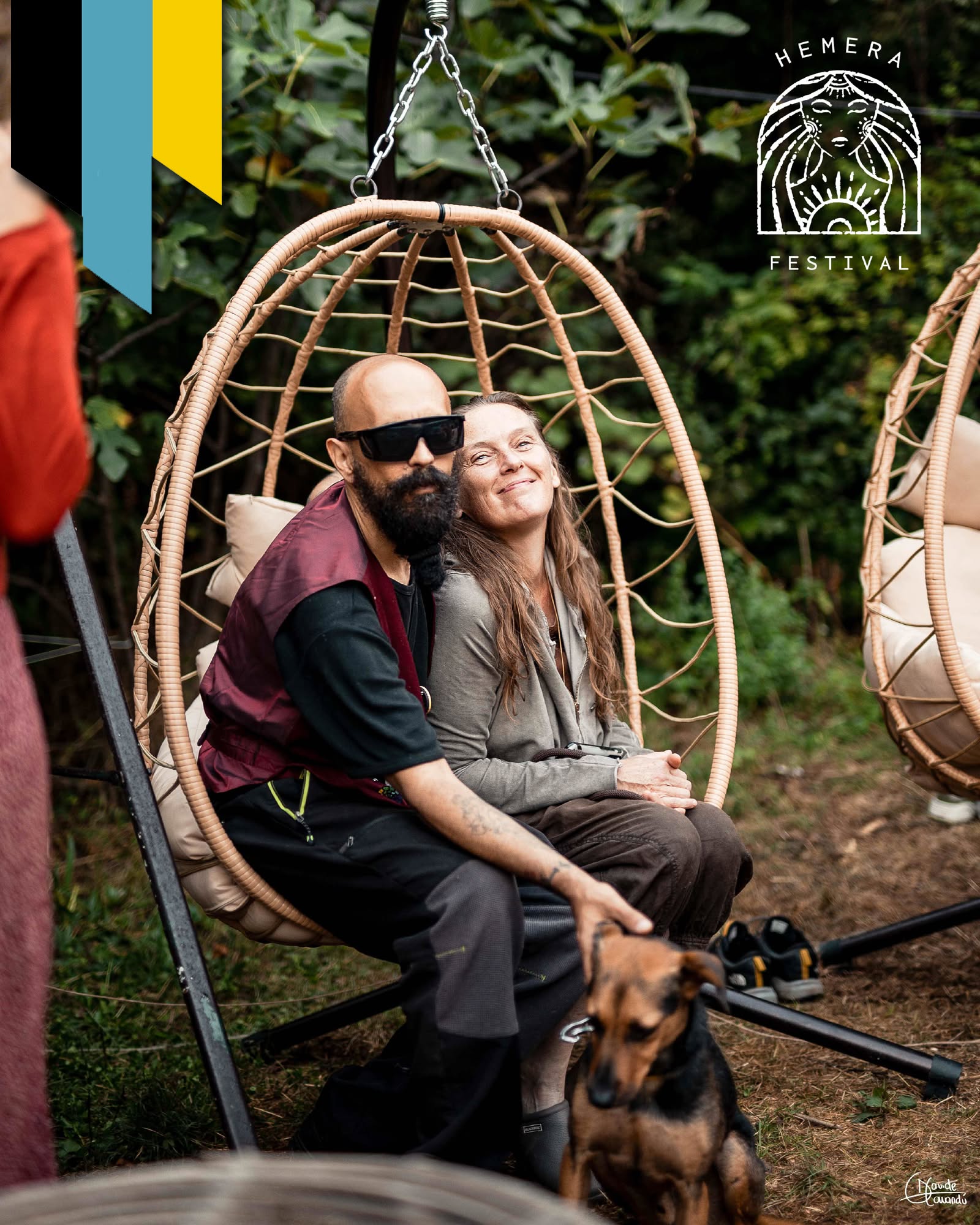 Two people sitting in a hanging chair outdoors, smiling, with a dog nearby, surrounded by greenery.