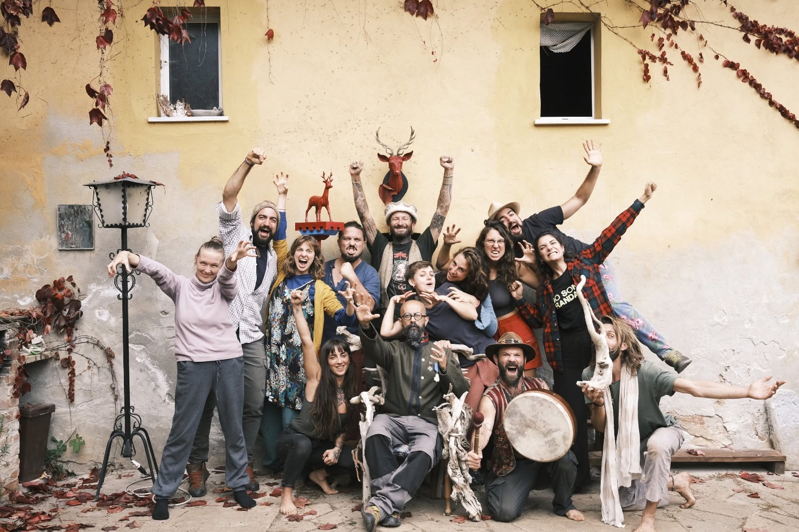 Group of people celebrating outdoors in front of a yellow wall, some raising hands, others playing drums, with a dog nearby.