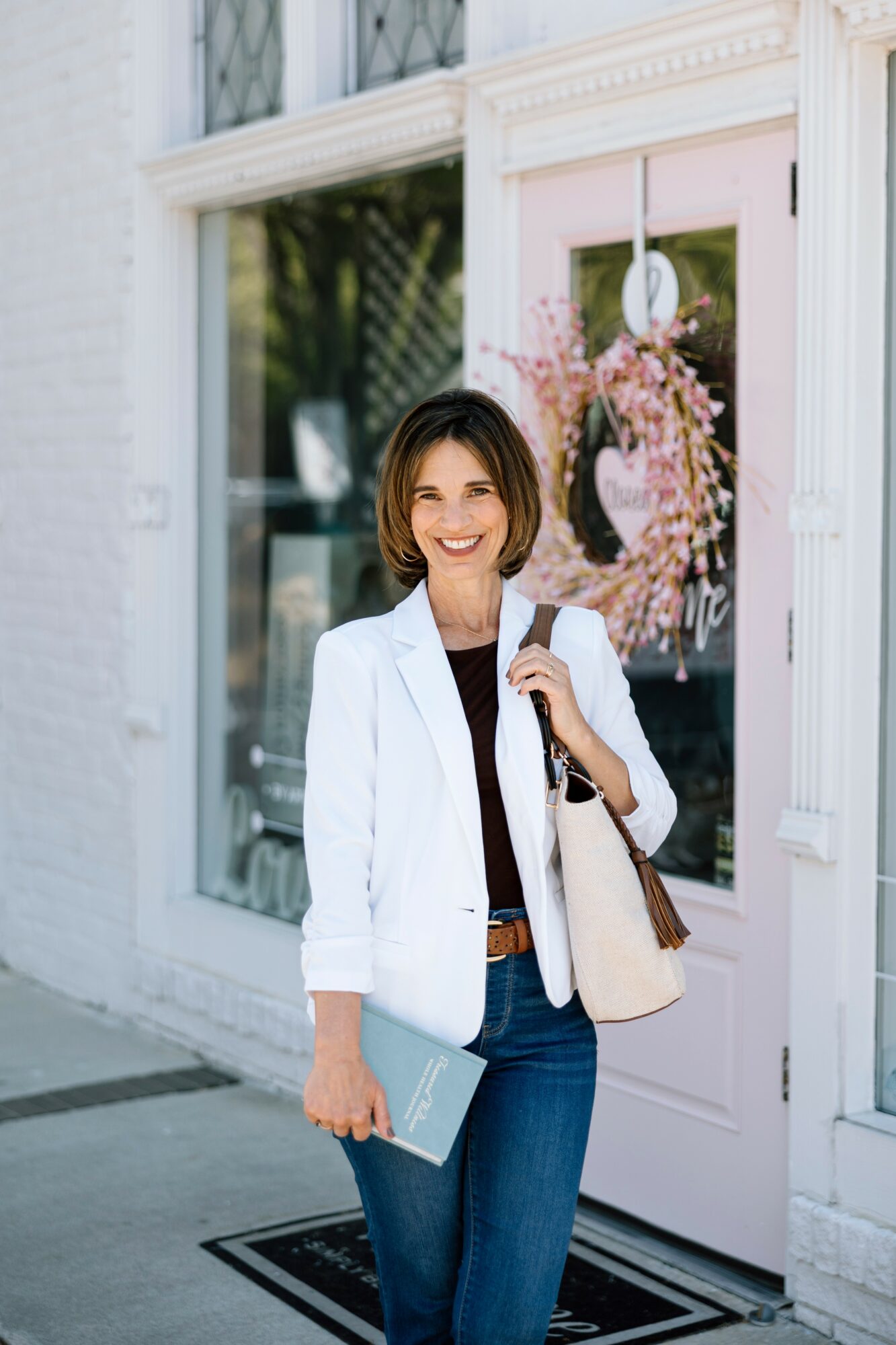 Woman standing outside a shop with pink door and floral wreath, smiling, holding a notebook and a bag.