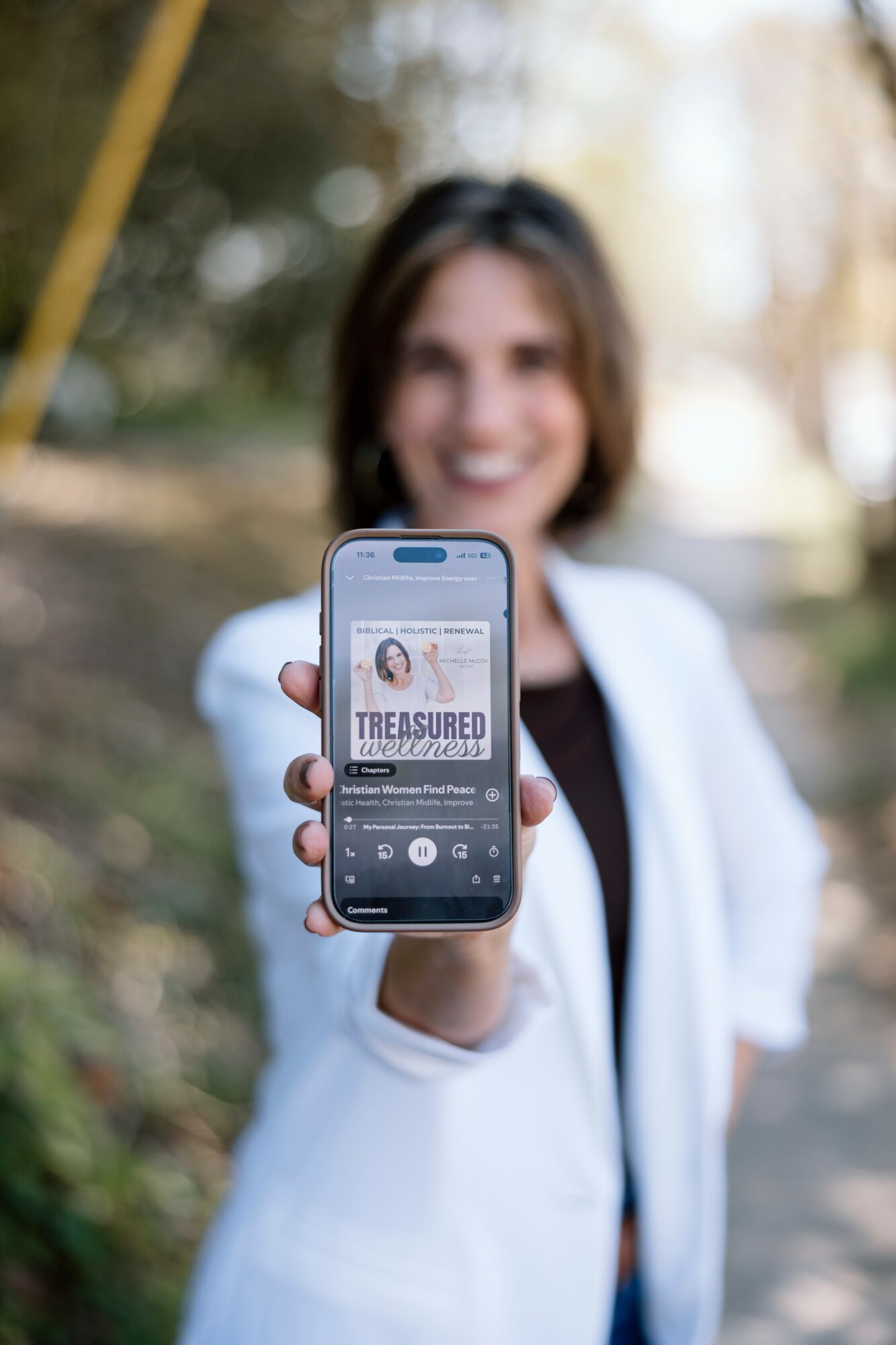 Person holding smartphone showing a music app with a smiling woman in the background outdoors.