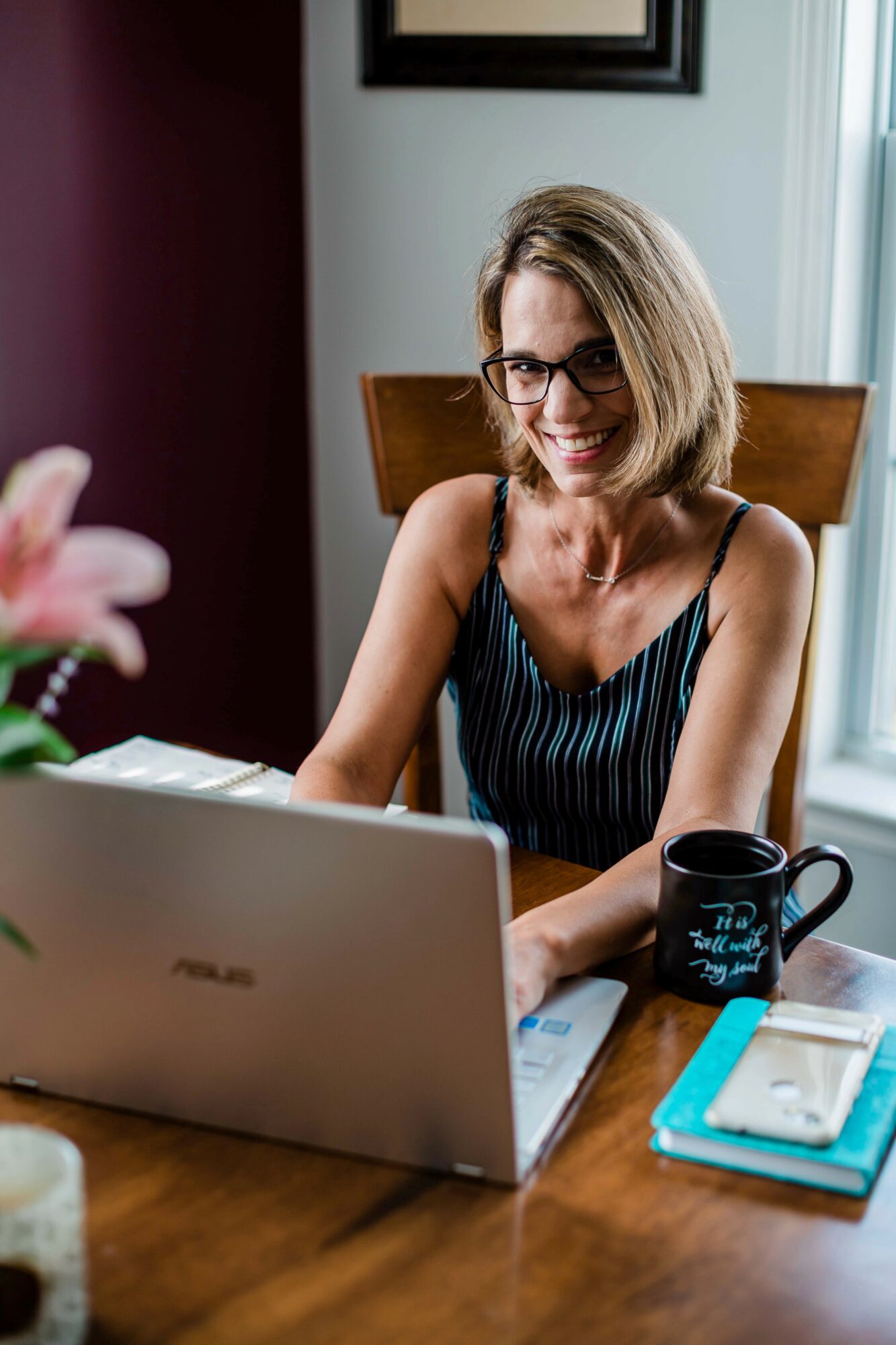 Woman with glasses working on a laptop at a wooden table, smiling, with a mug and notebook nearby.