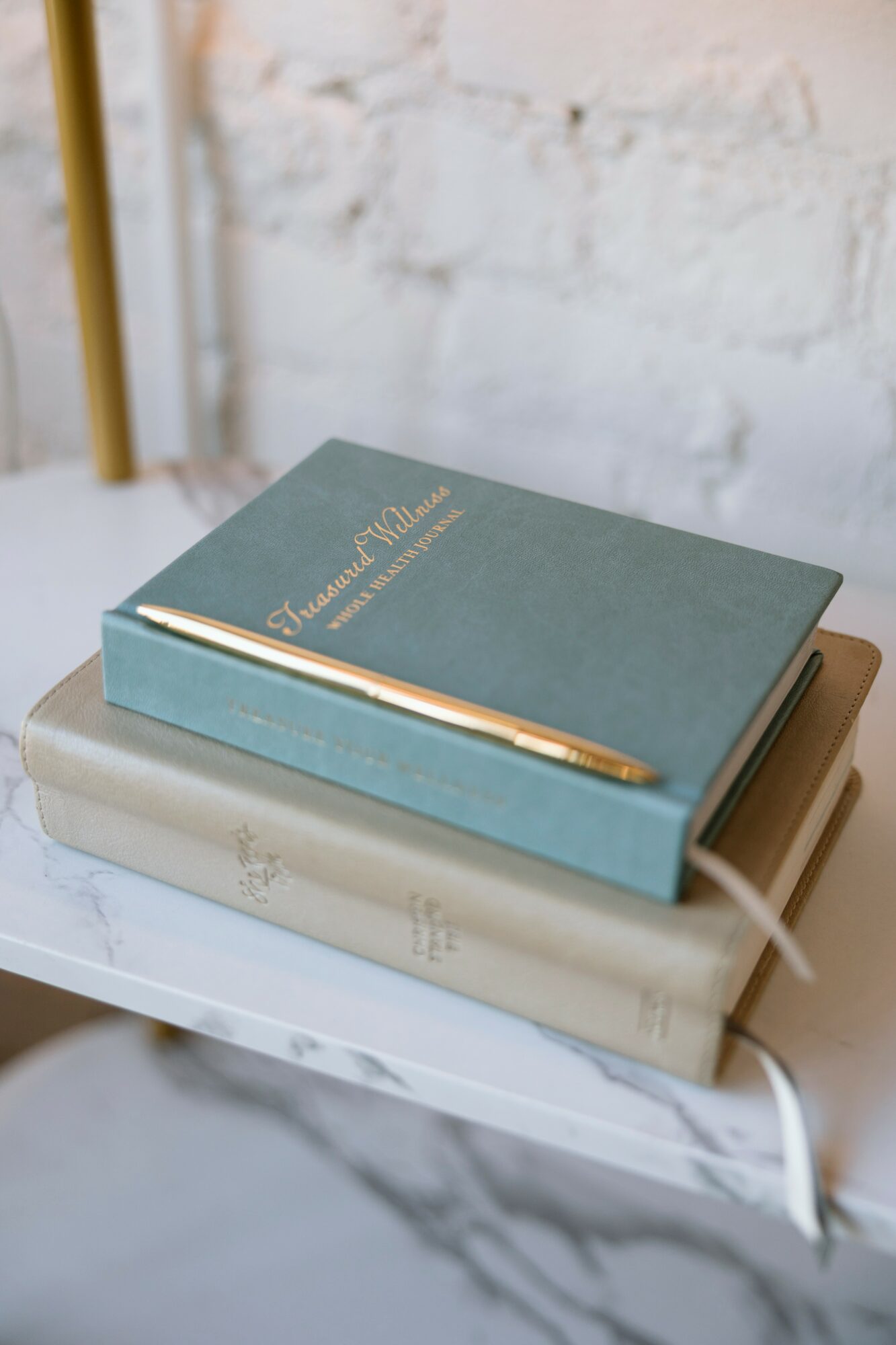 Two closed books stacked on a white marble surface, with a gold pen resting on top of the upper book, against a white brick wall background.