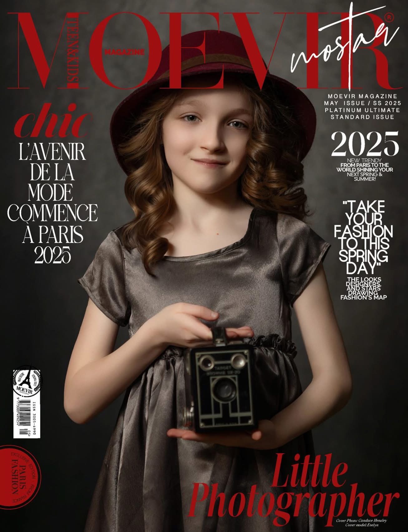 Young girl with wavy brown hair holding a vintage camera, wearing a shiny gray dress, smiling softly.