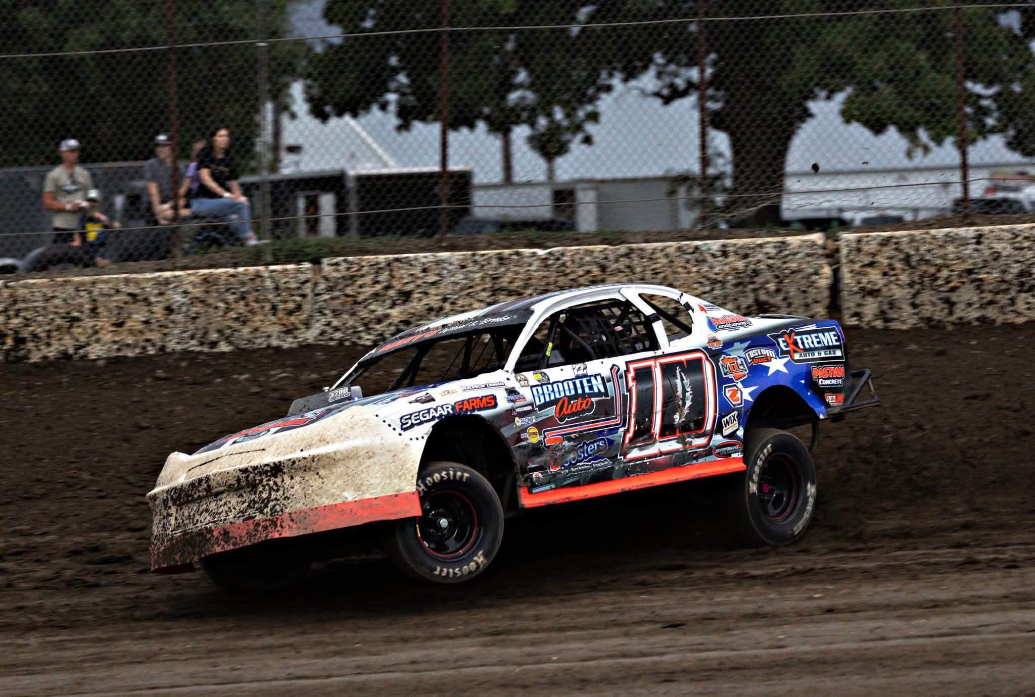 Race car on dirt track with spectators and trees in background.