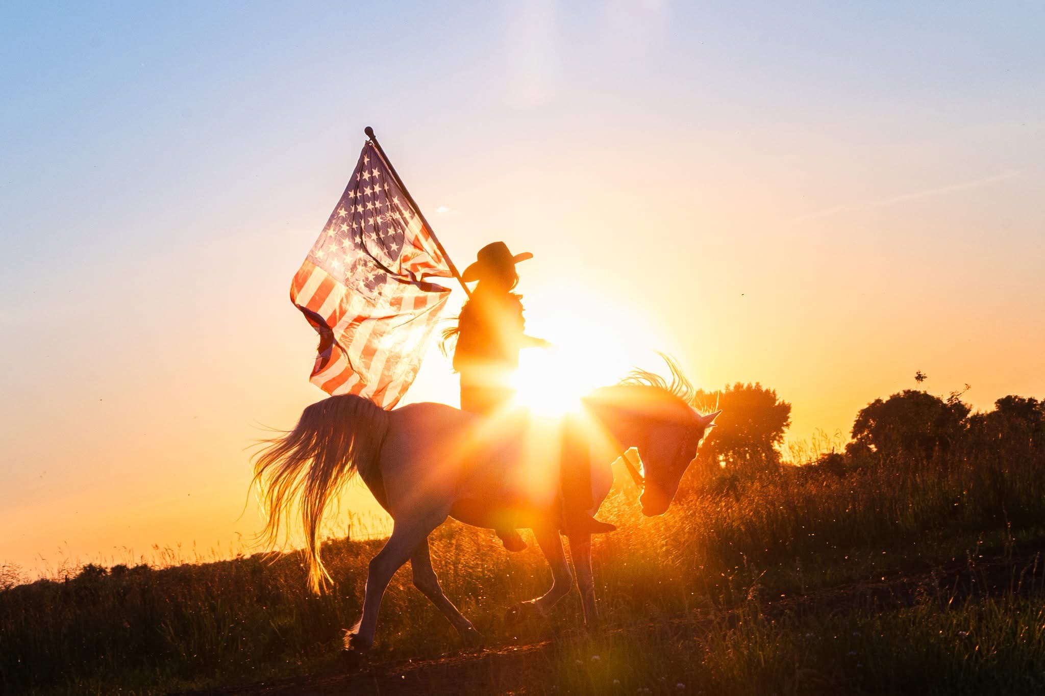 Person riding a horse holding an American flag during sunset, with trees in the background.