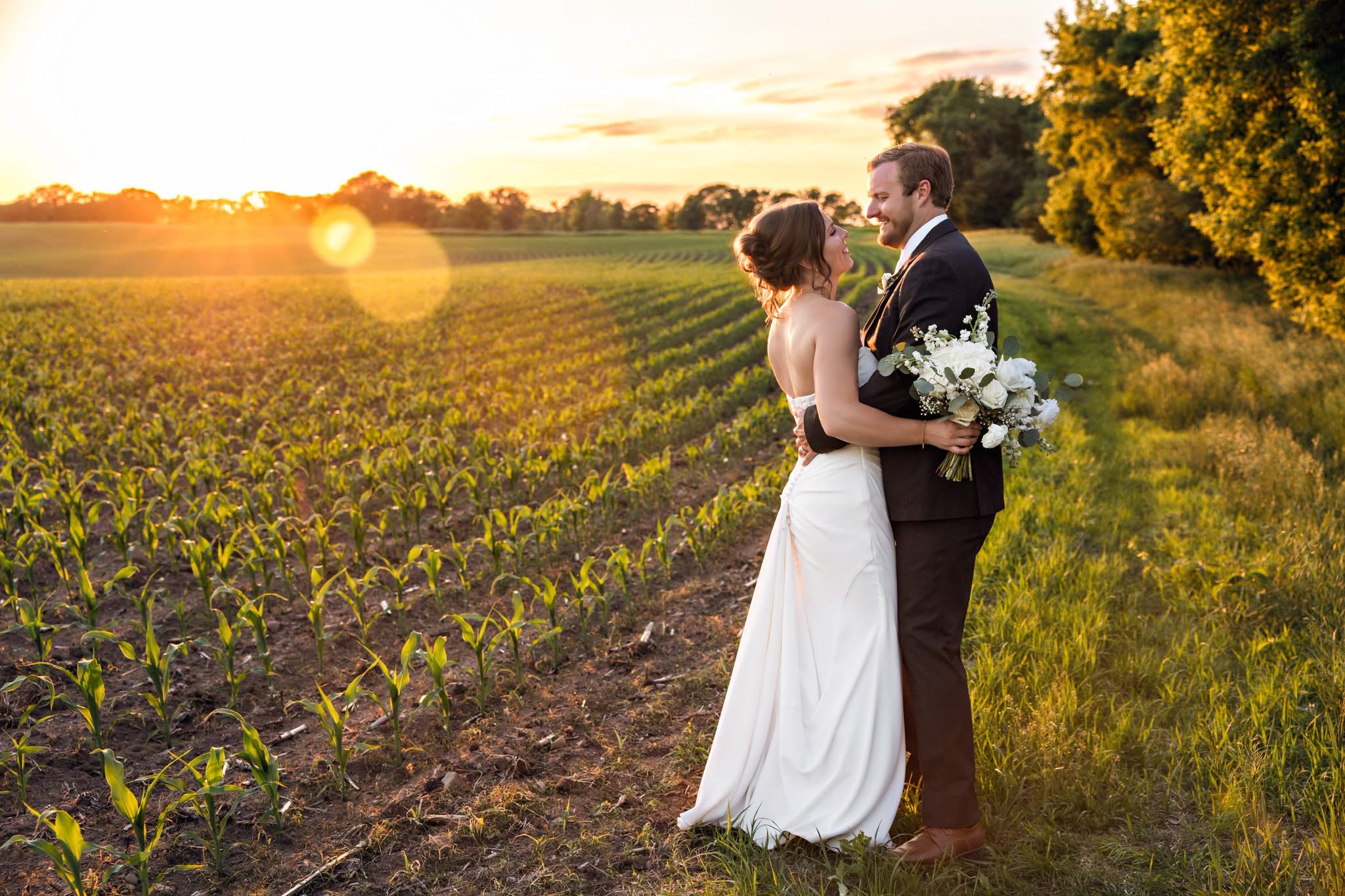 A bride and groom stand in a field at sunset, holding a bouquet, facing each other, with trees in the background.