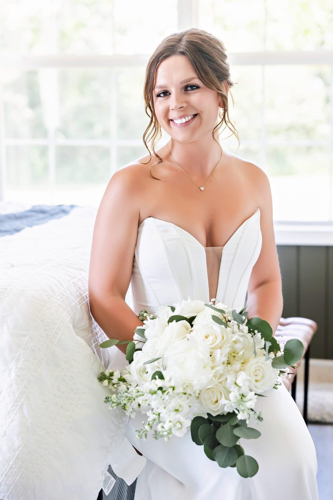 Smiling woman in wedding dress holding a bouquet of white flowers, sitting indoors near a window with natural light.