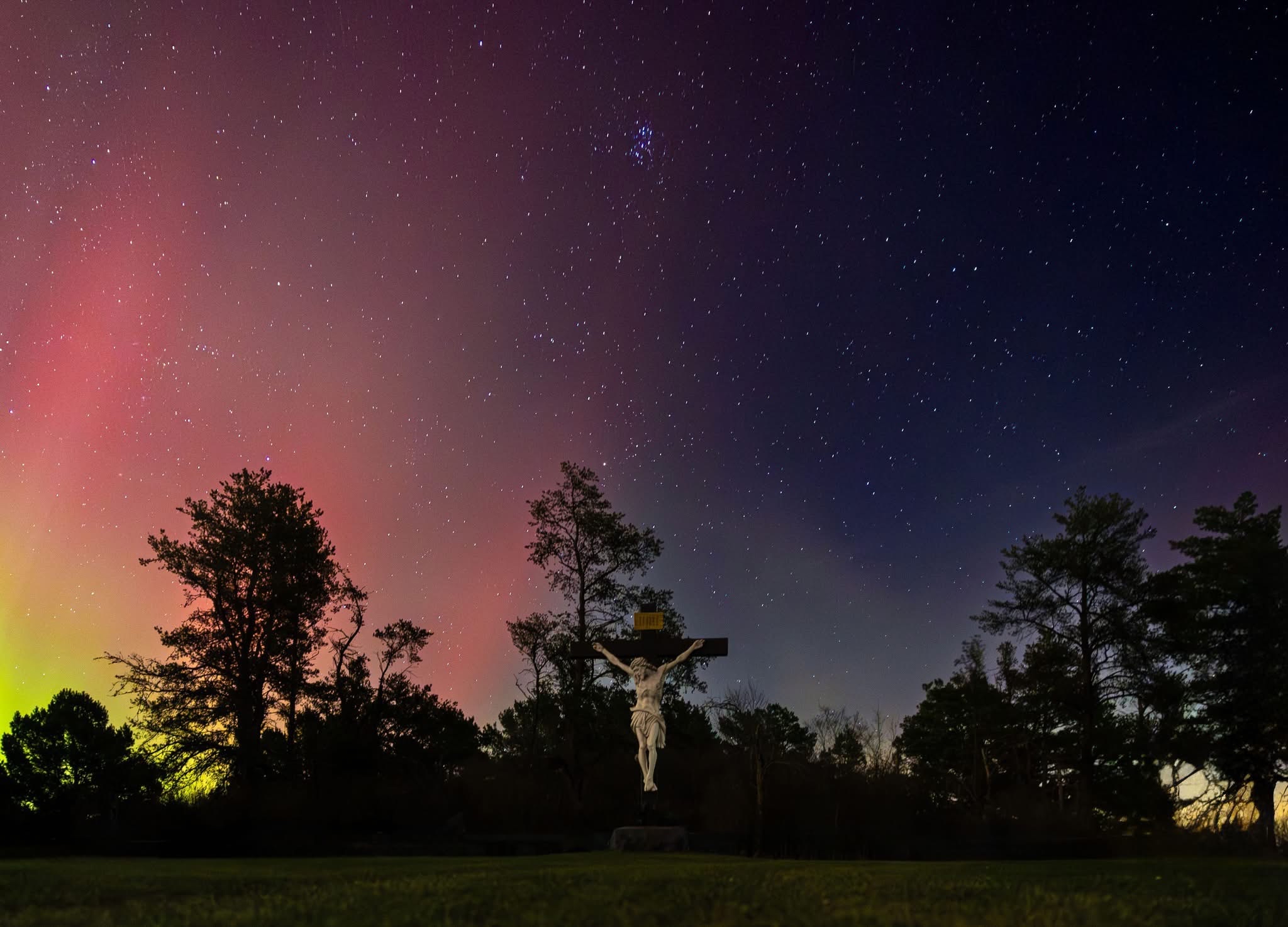 Night sky with stars and colorful aurora above trees and a crucifix in the foreground.