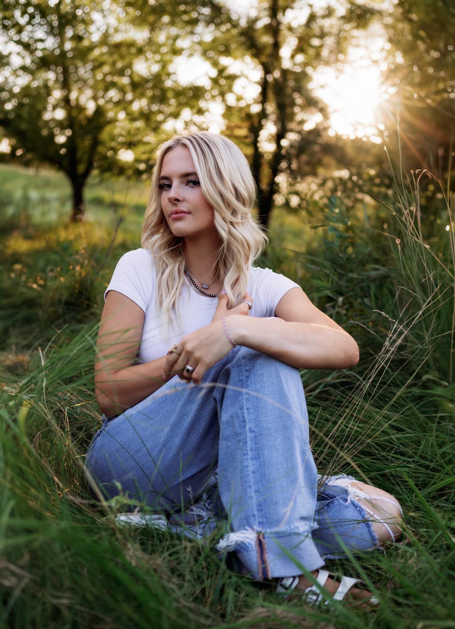 Young woman with blonde hair sitting on grass in a park during sunset, wearing a white shirt and jeans.