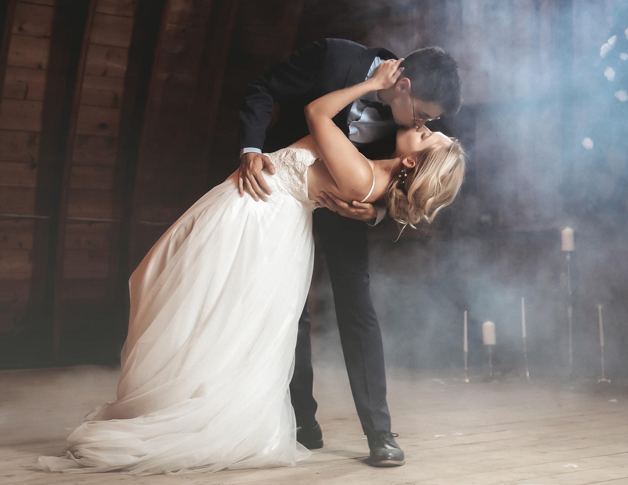 A couple dancing closely, with the man in a suit and the woman in a white dress, in a dimly lit setting.