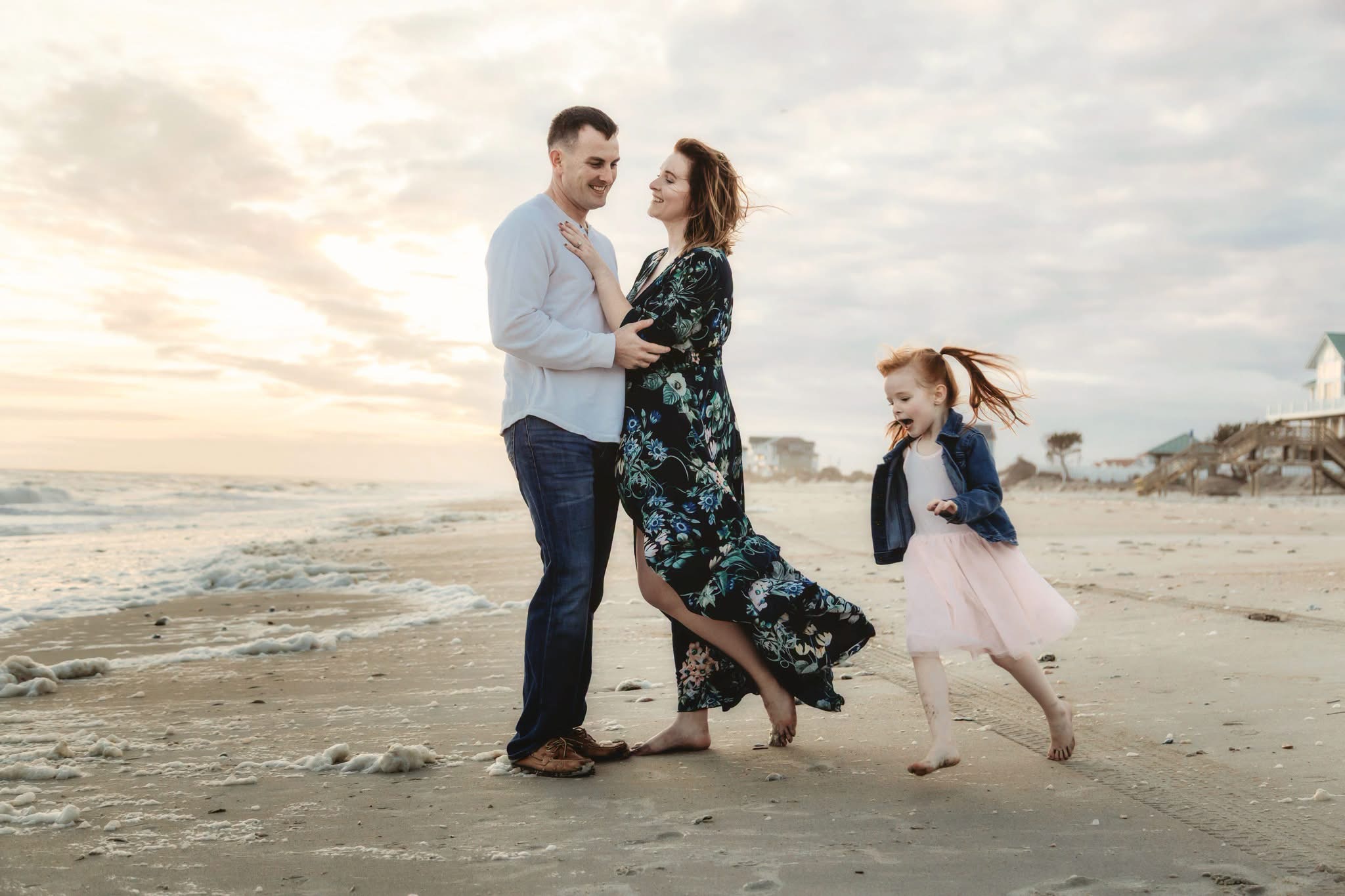 Family of three on the beach, a man and woman holding hands, a girl running nearby, sunset in background