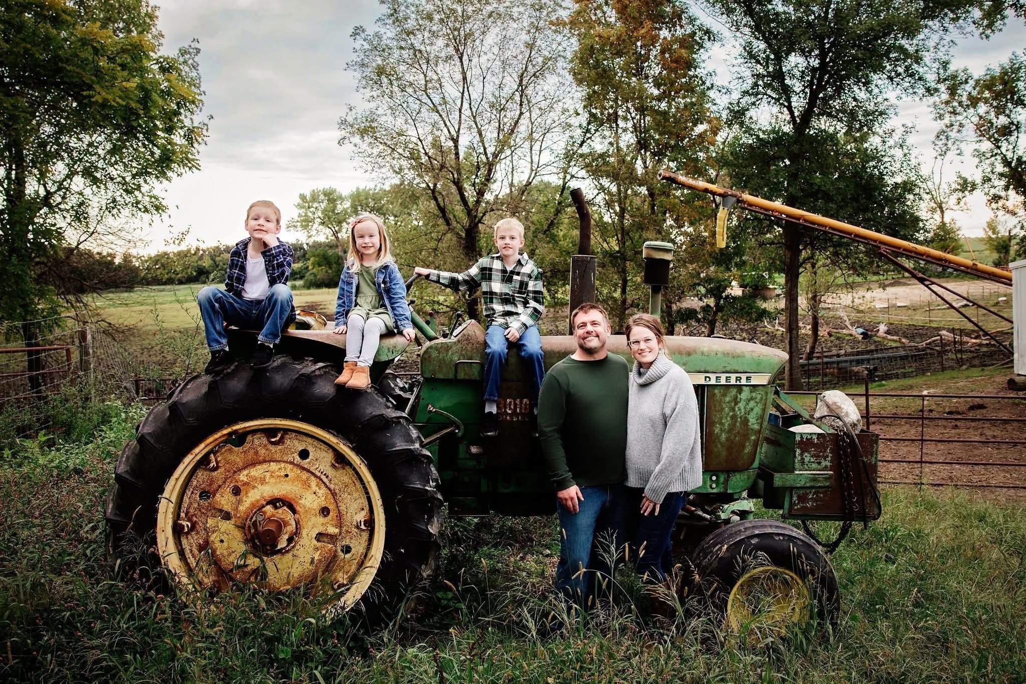 Four children and two adults on and beside a green tractor outdoors with trees and a fence in the background.