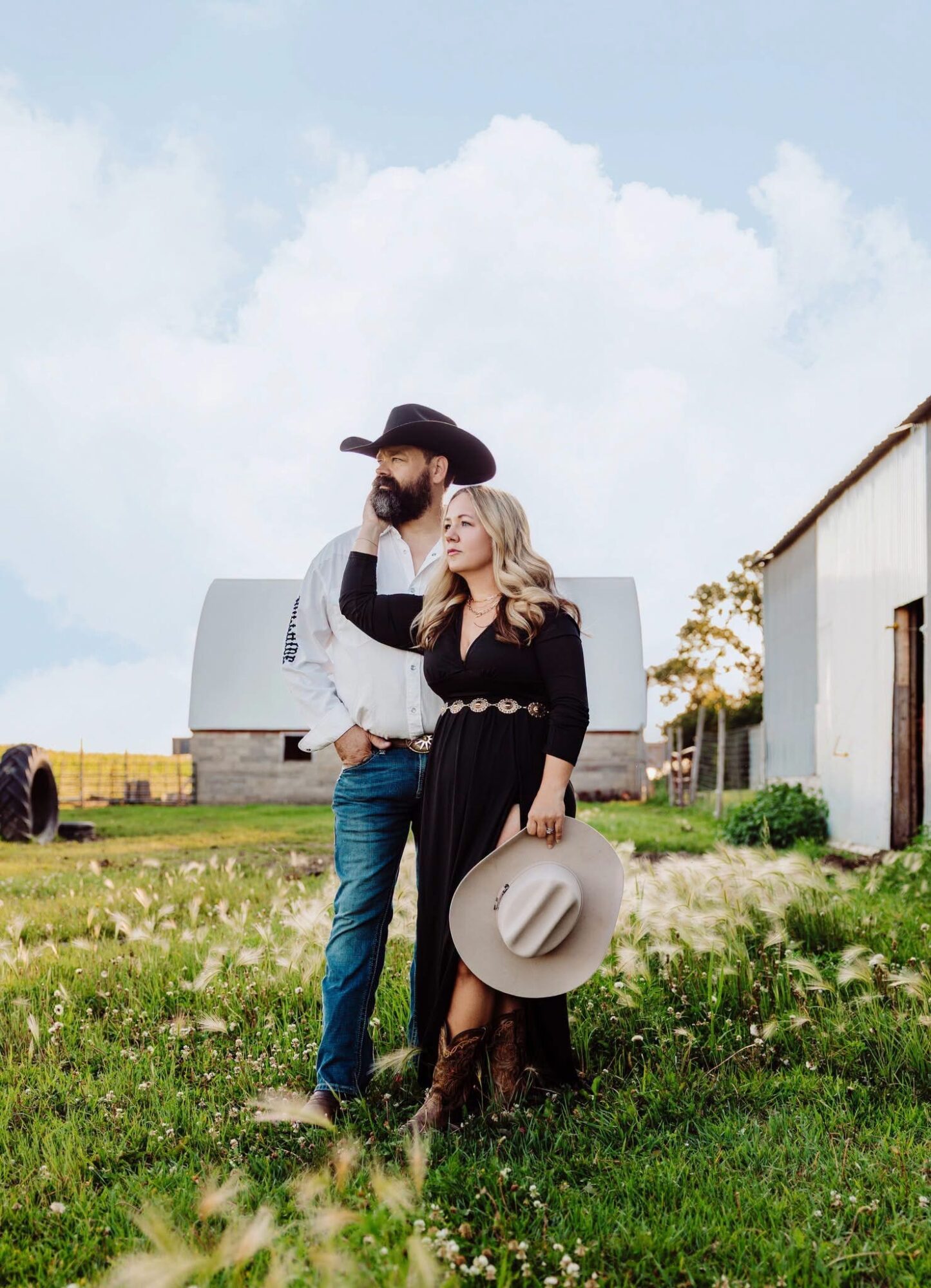 A man and woman stand outdoors in front of farm buildings, with the woman holding a large hat, both dressed in vintage style.