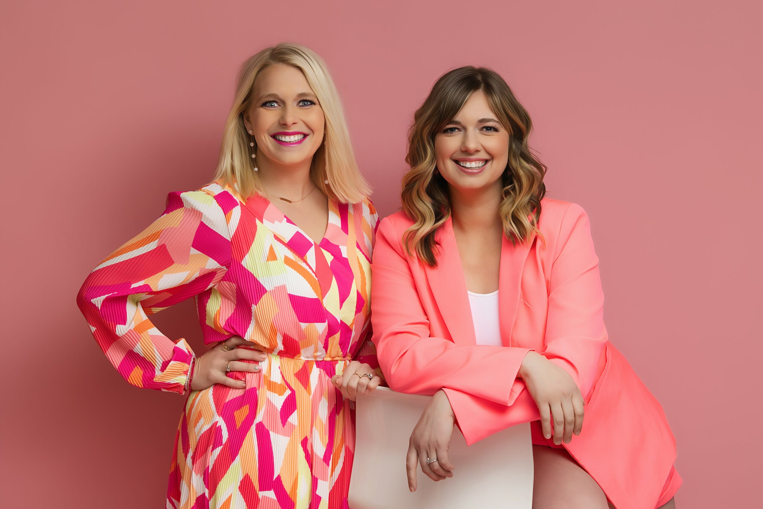 Two women smiling, one in a colorful patterned dress, the other in a pink blazer, standing against a pink background.