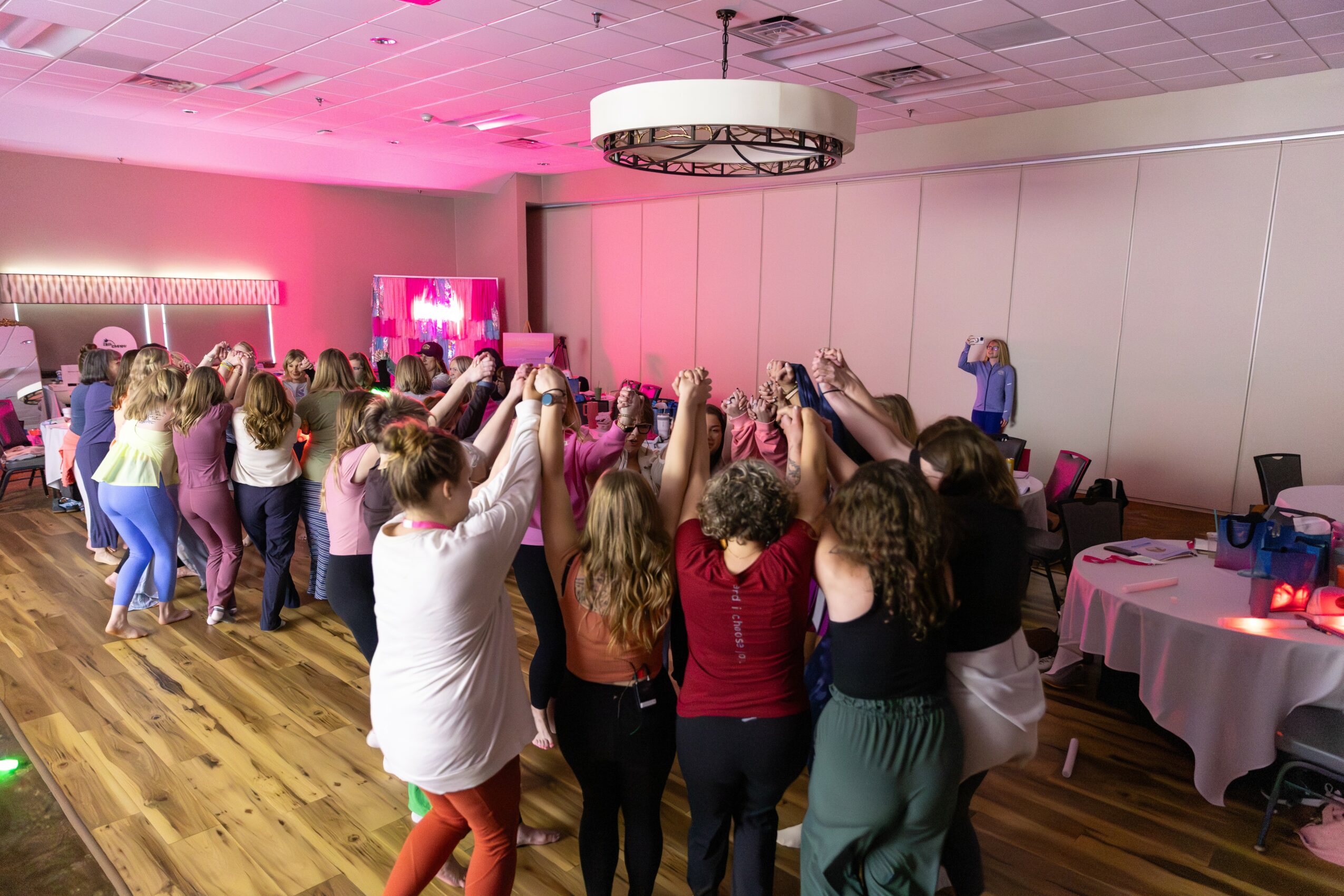 Group of people dancing in a room with pink lighting, some raising hands, with tables on the side.