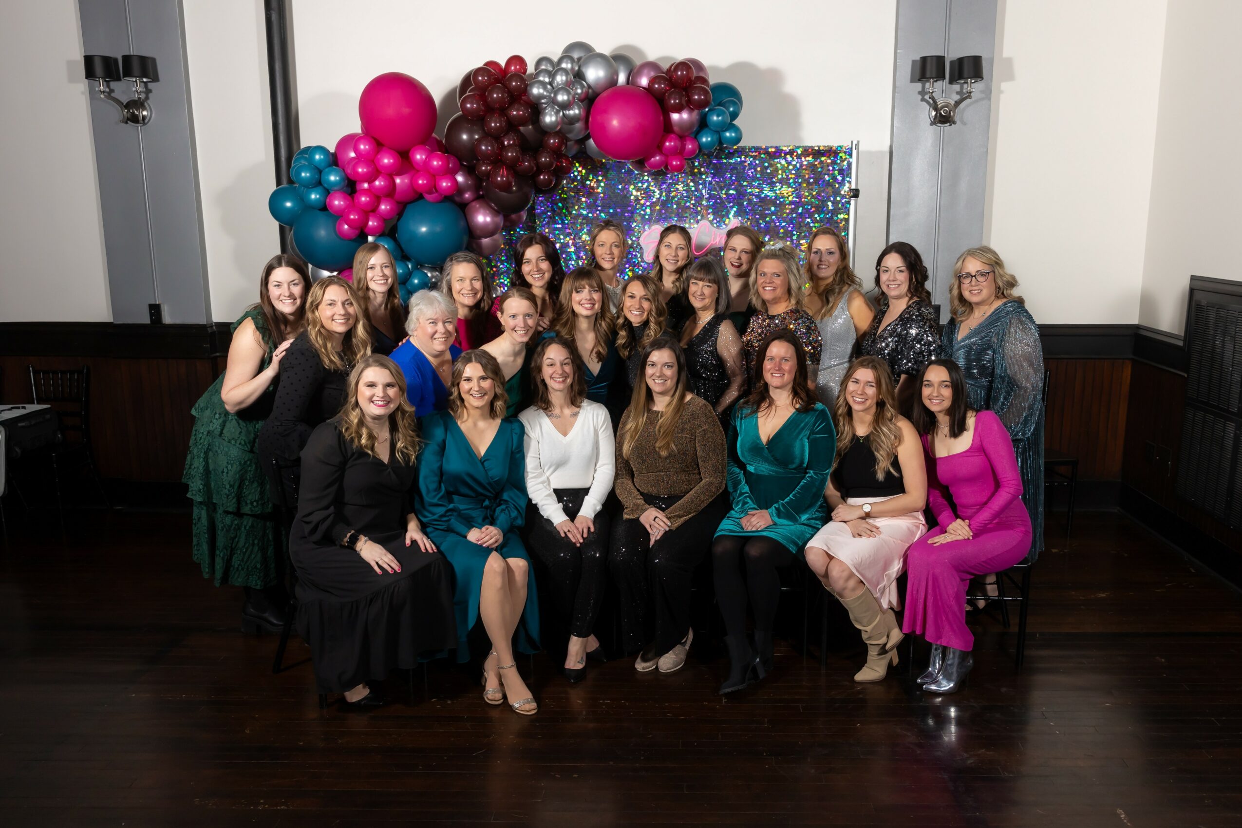 Group of women posing in front of colorful balloon decorations, smiling, indoors with dark wood floors and white walls.