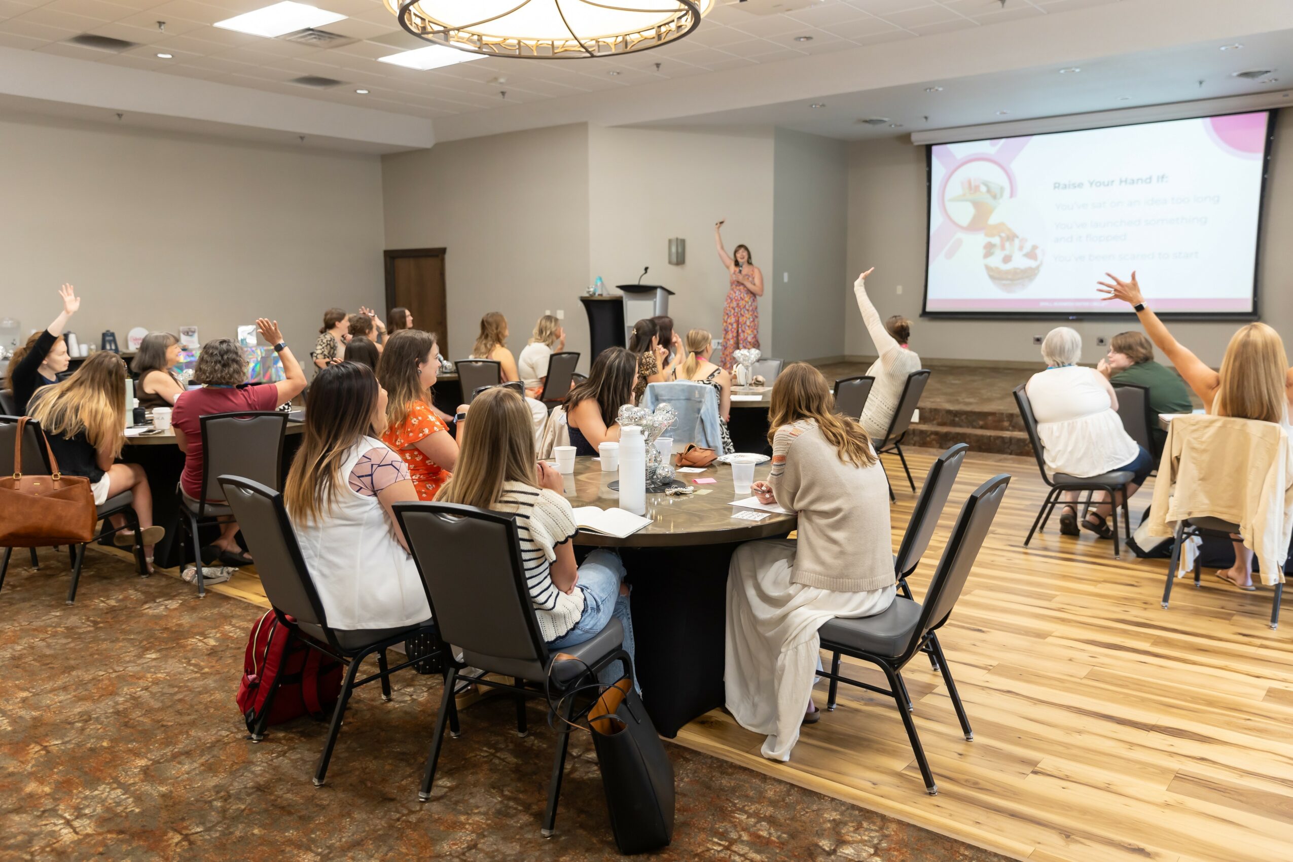 Conference room with people seated at tables, some raising hands, presenter at front with screen, wooden floor, ceiling light fixture.
