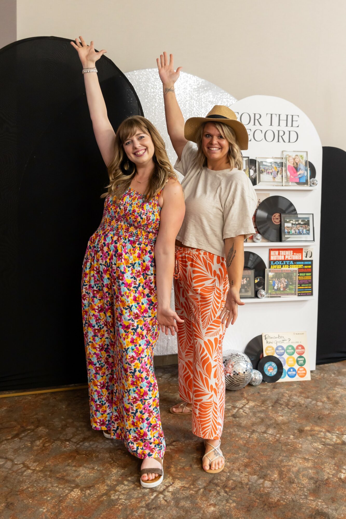 Two women stand in front of a display, raising their arms and smiling, one wearing a colorful jumpsuit and the other in a beige top and patterned pants.