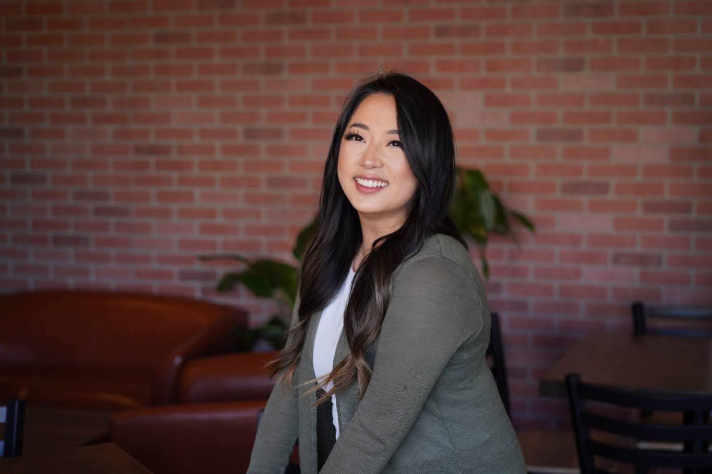 Smiling woman with long dark hair in a gray blazer and white top, sitting indoors with a brick wall background.