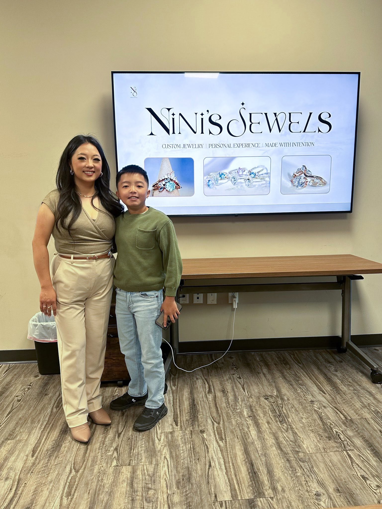 Woman and boy standing in front of a large screen displaying jewelry images, smiling, in a room with beige walls and wooden floor.