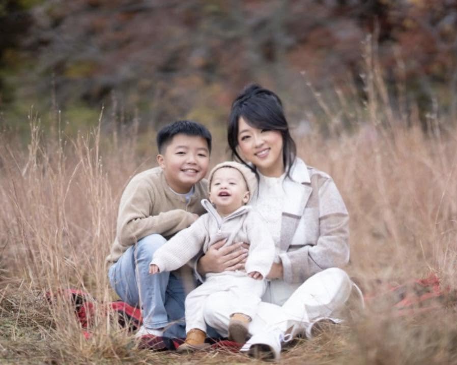 Family of three sitting outdoors in tall grass, smiling, with autumn trees in background.