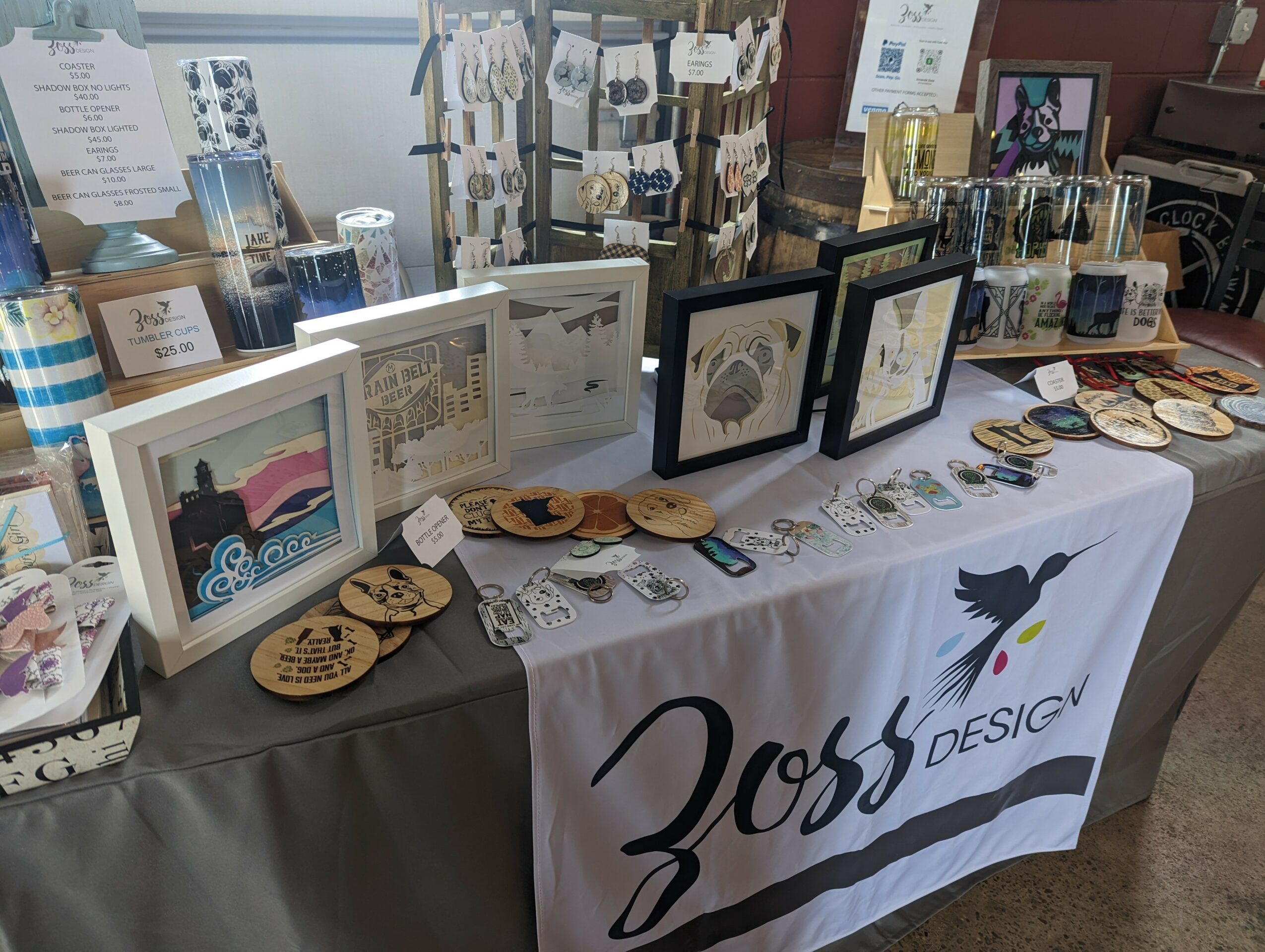 Display table with framed artwork, wooden items, jewelry, and a banner reading 'Doss Design' in a shop or market setting.