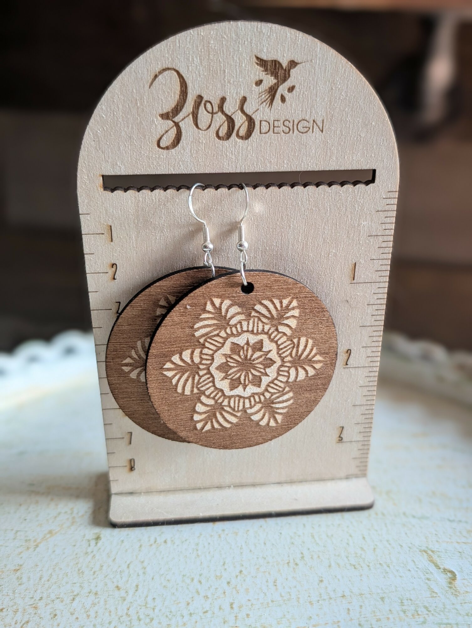 Wooden display with a circular tag hanging, featuring a floral design, and the text 'Zoss DESIGN' at the top.