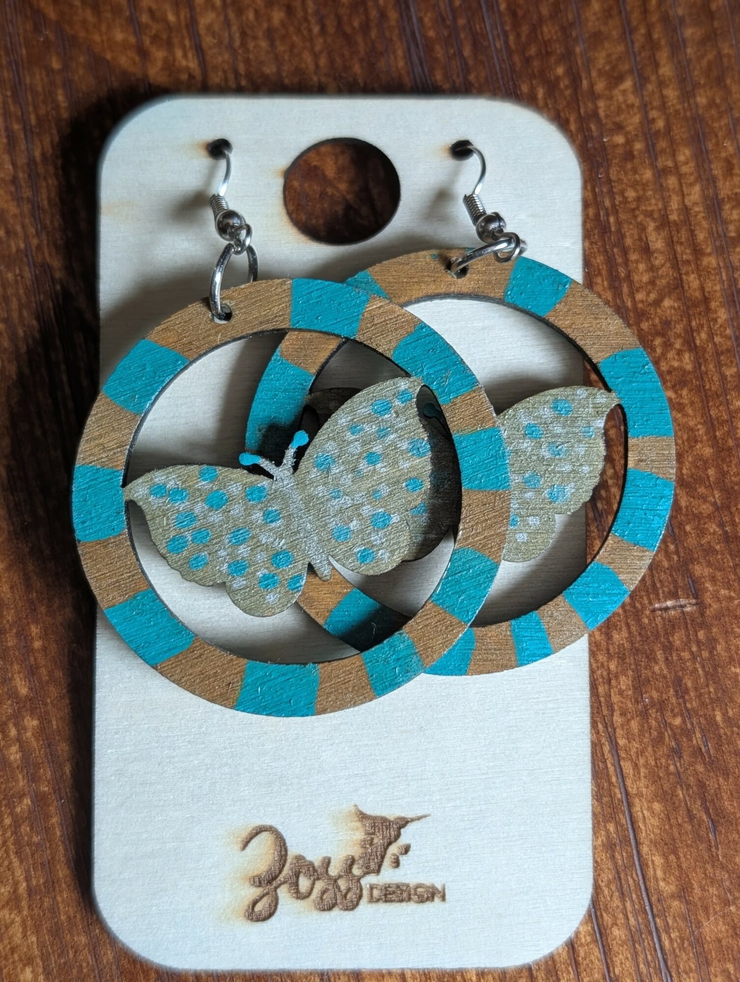 Colorful butterfly earrings with circular hoops on a white card, placed on a wooden surface.