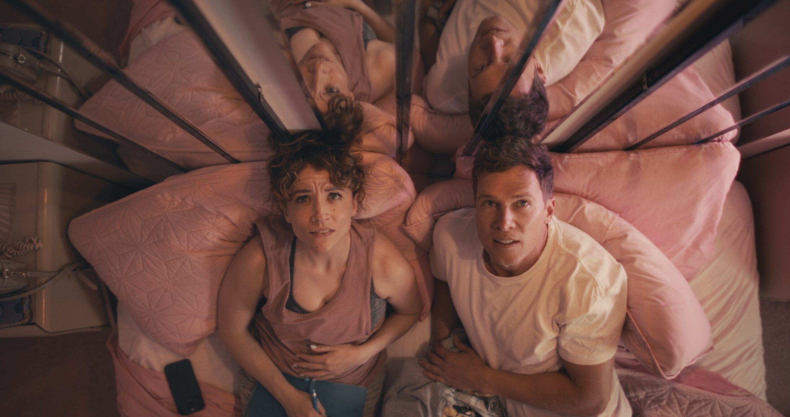 Two people sit on a bed with pink bedding, looking up at the camera, which is above them. Their reflections are visible in a mirror behind them.