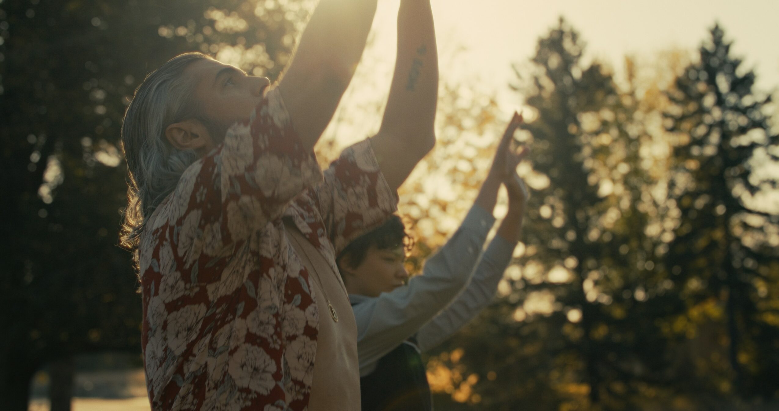 Two people outdoors with trees in the background, raising their arms in the sunlight.