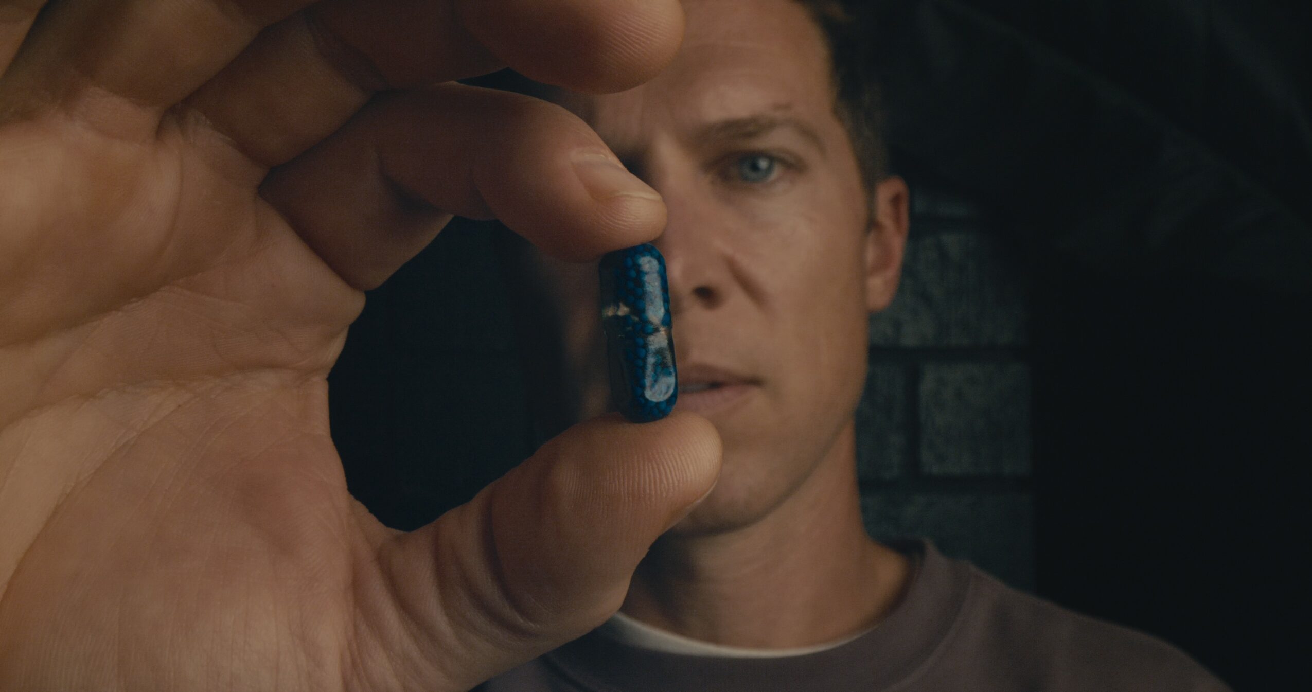 Person holds a small blue object close to the camera, with a serious expression, dark background, and brick wall.