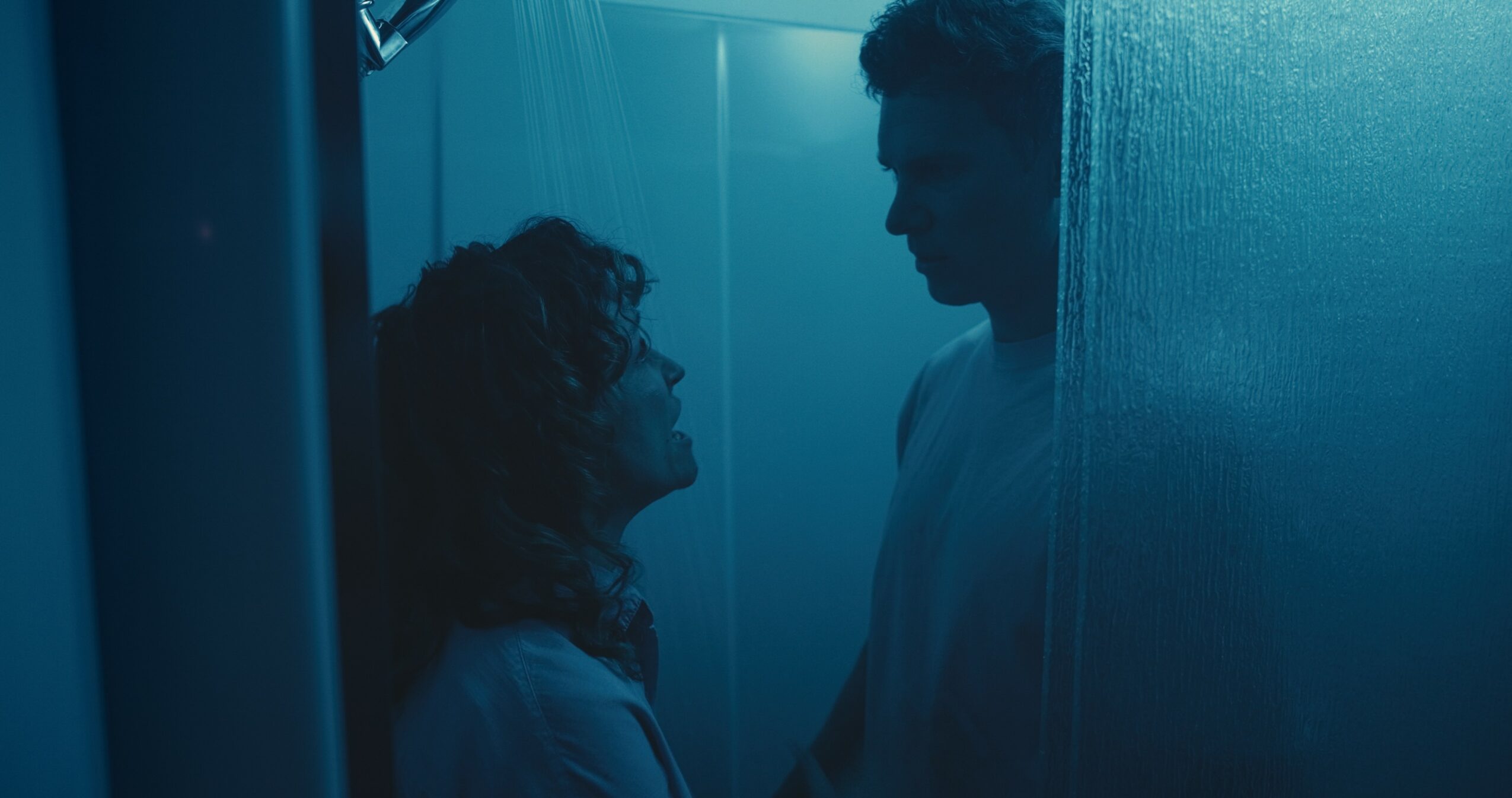Two people face each other in a dimly lit, blue-toned room, standing close and looking at each other.