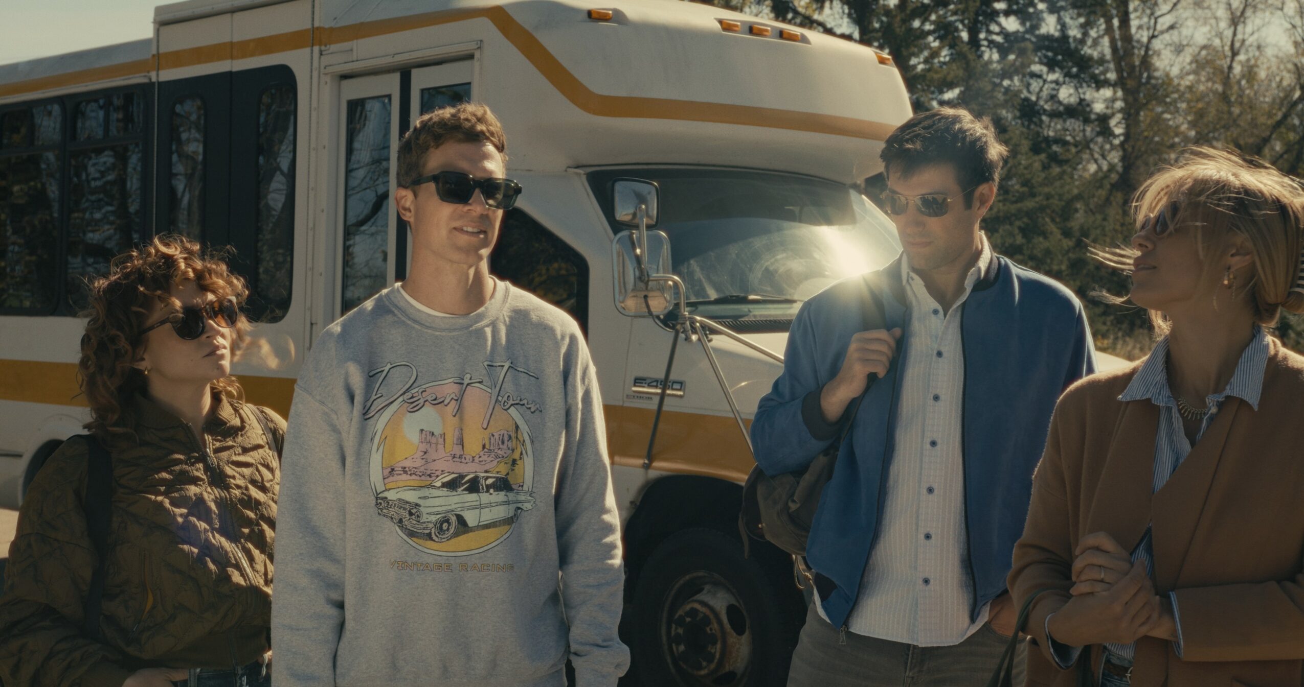 Four people standing outdoors near a large vehicle, with trees in the background.