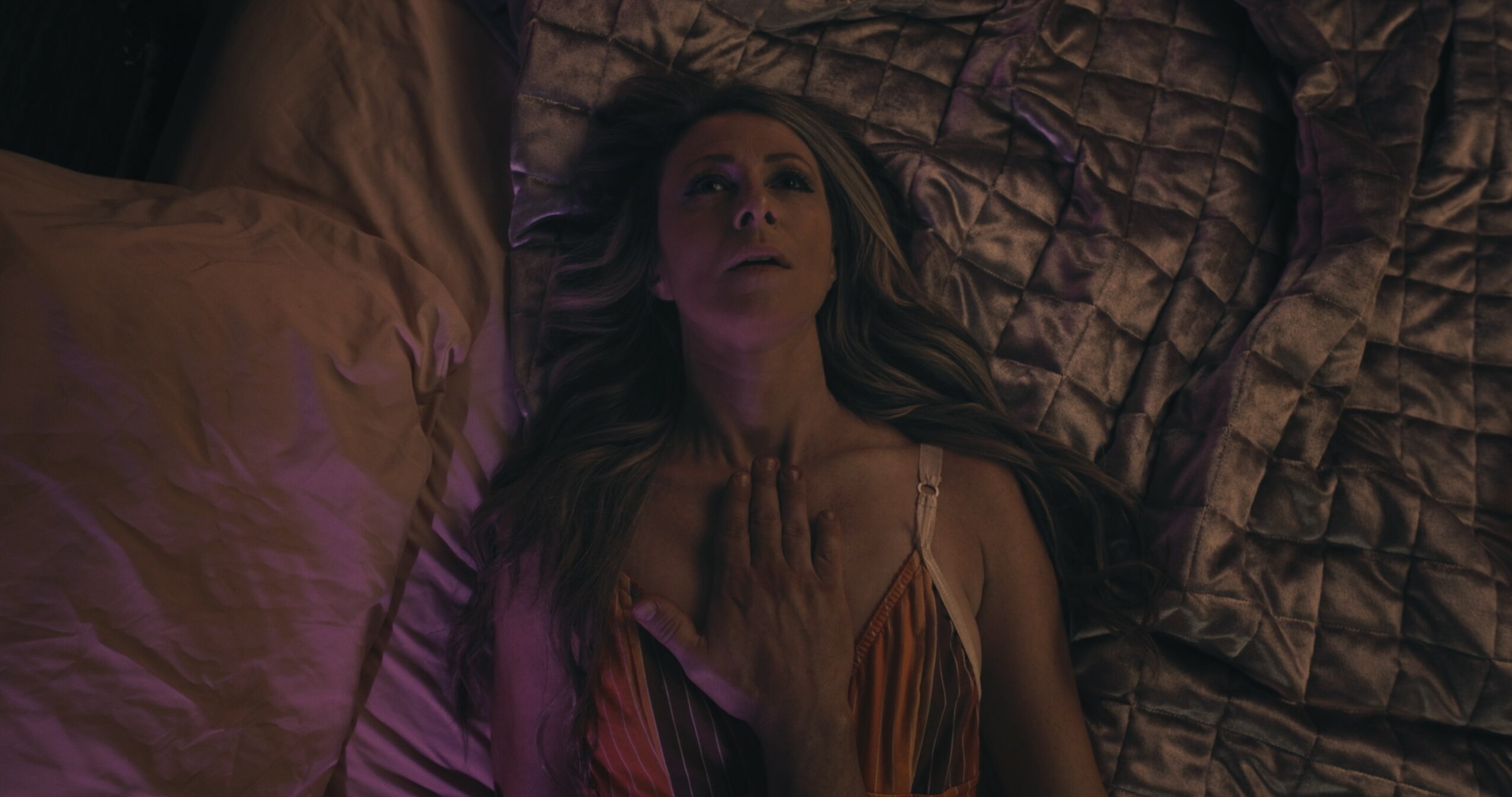 Woman lying on bed with purple and brown bedding, looking upward, with long hair and a colorful dress.