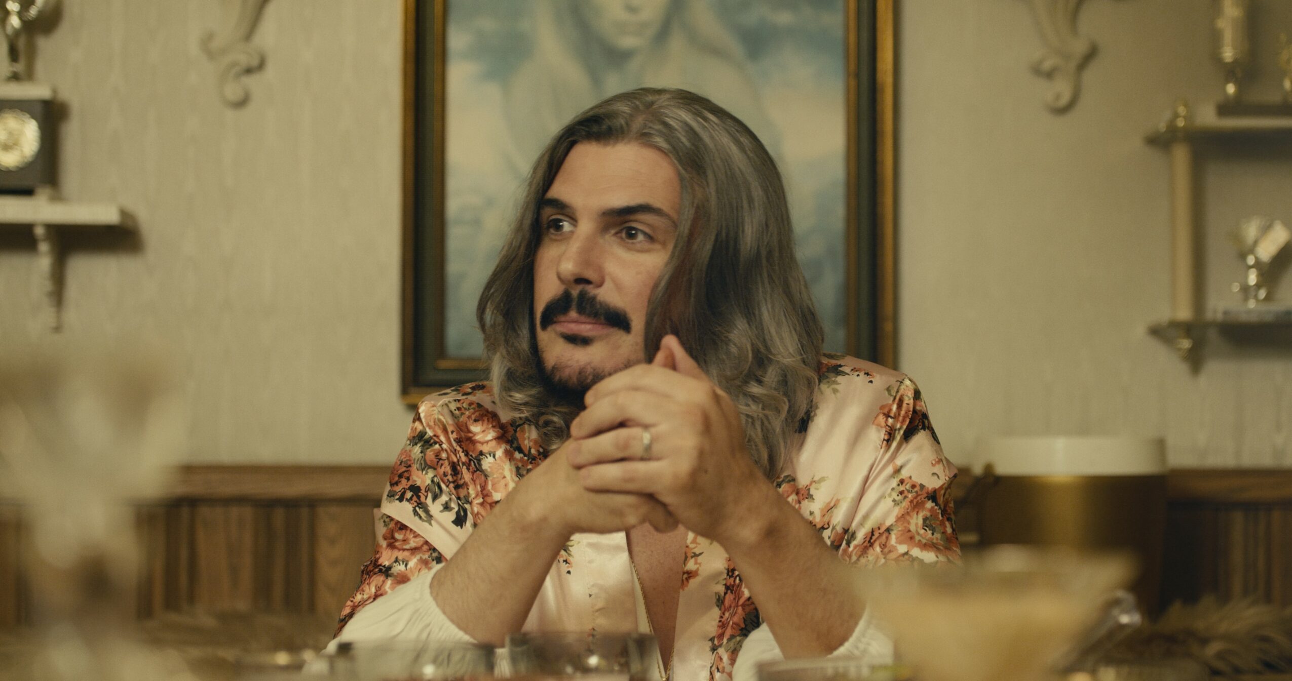 Man with long hair and beard sitting at a table, holding his hands together, in a room with shelves and a framed picture behind him.