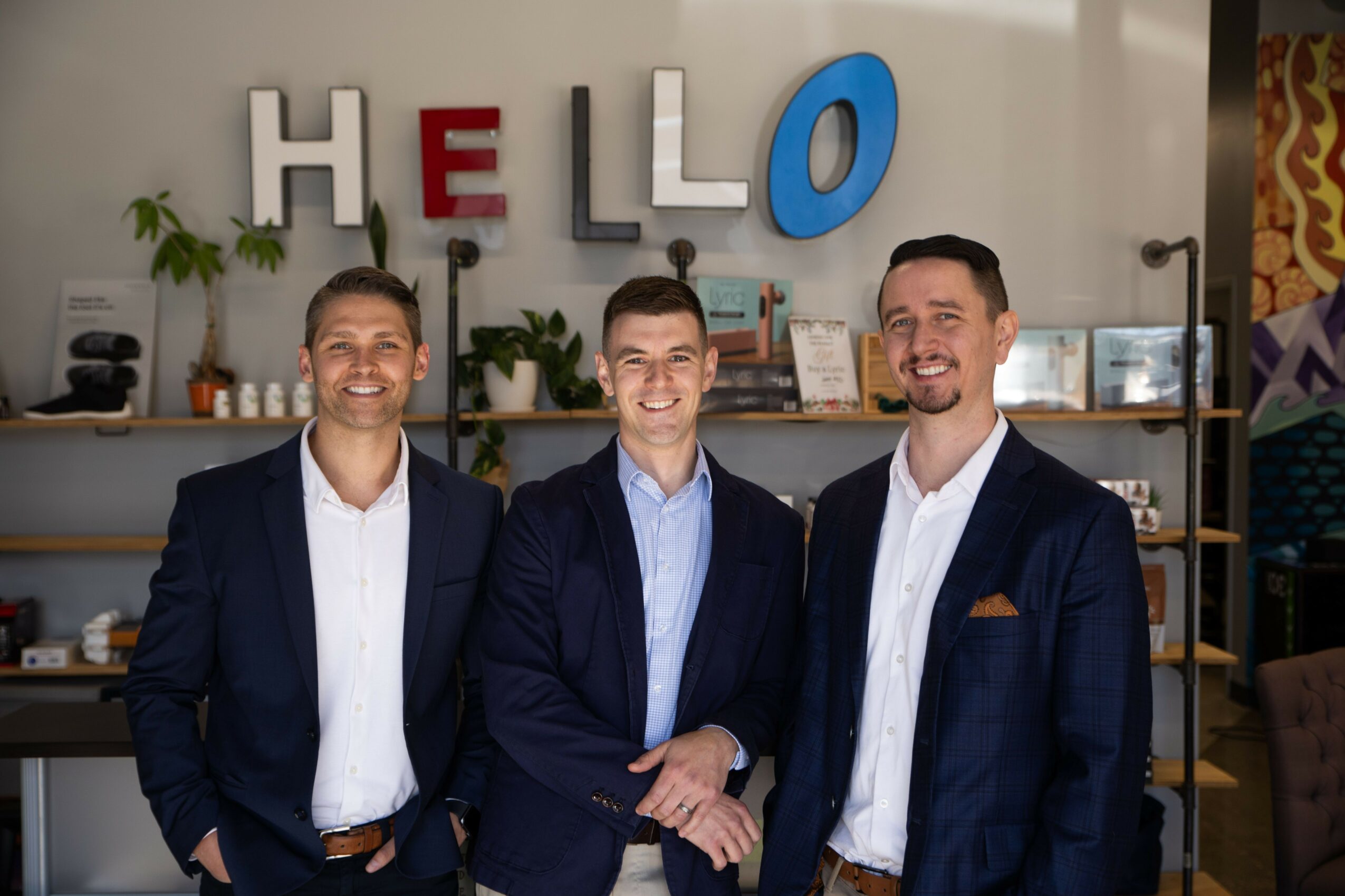 Three men in suits standing in front of a wall with colorful letters spelling 'HELLO'.