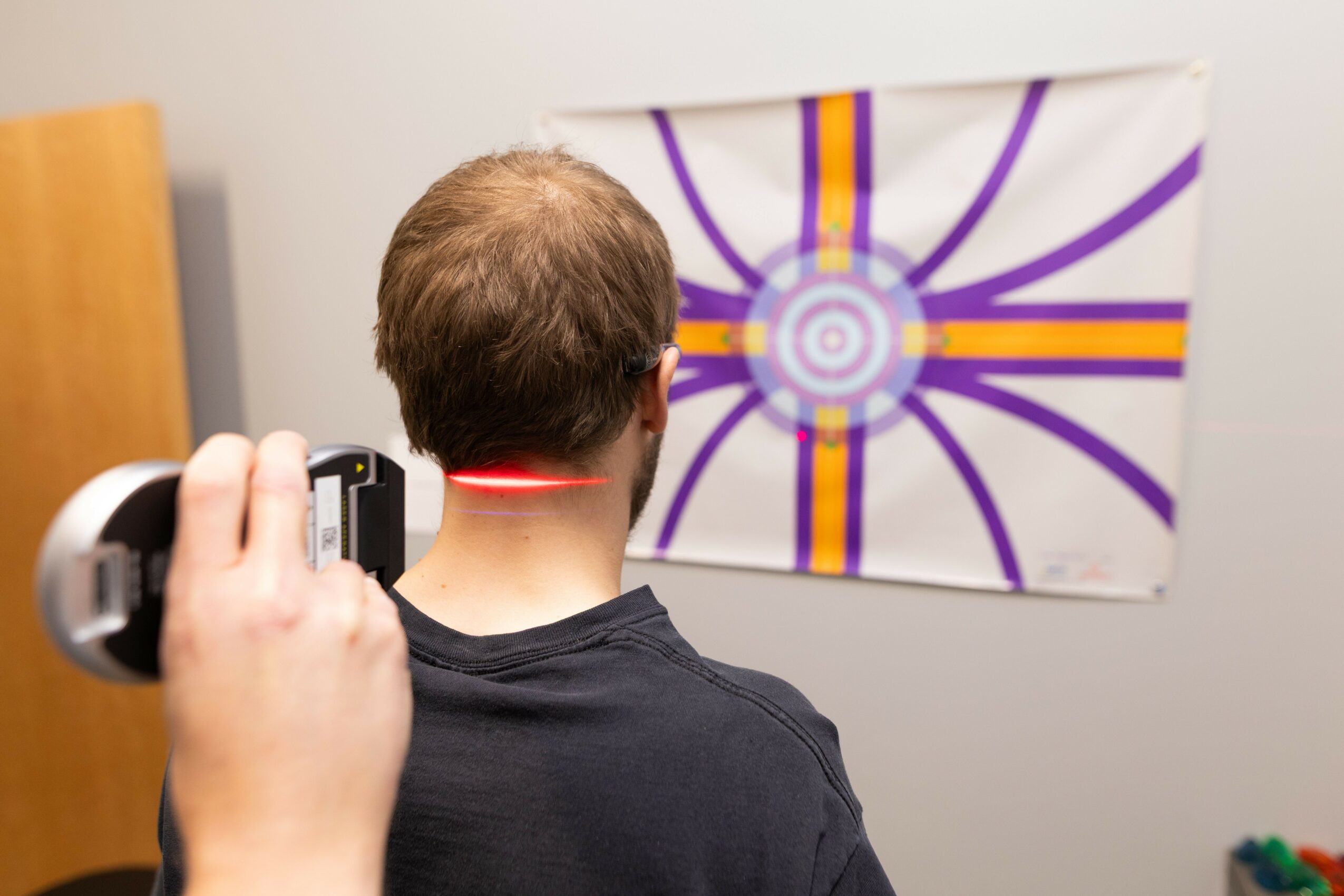 Person with short hair and black shirt using a handheld device to scan a red laser on the back of their neck, facing a colorful abstract wall art.