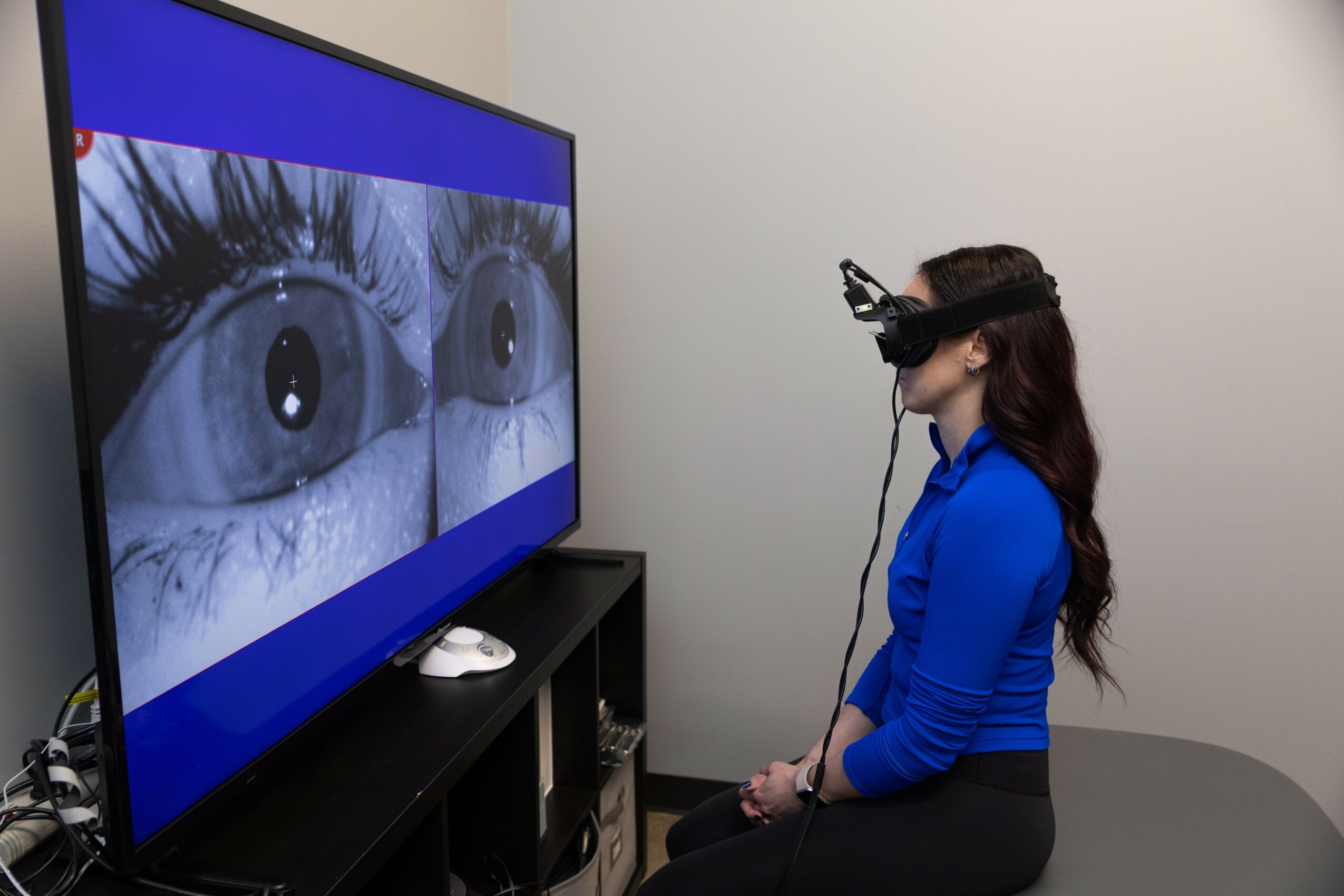 Woman wearing a virtual reality headset, sitting in front of a large screen displaying a close-up of eyes, in a room with plain walls.