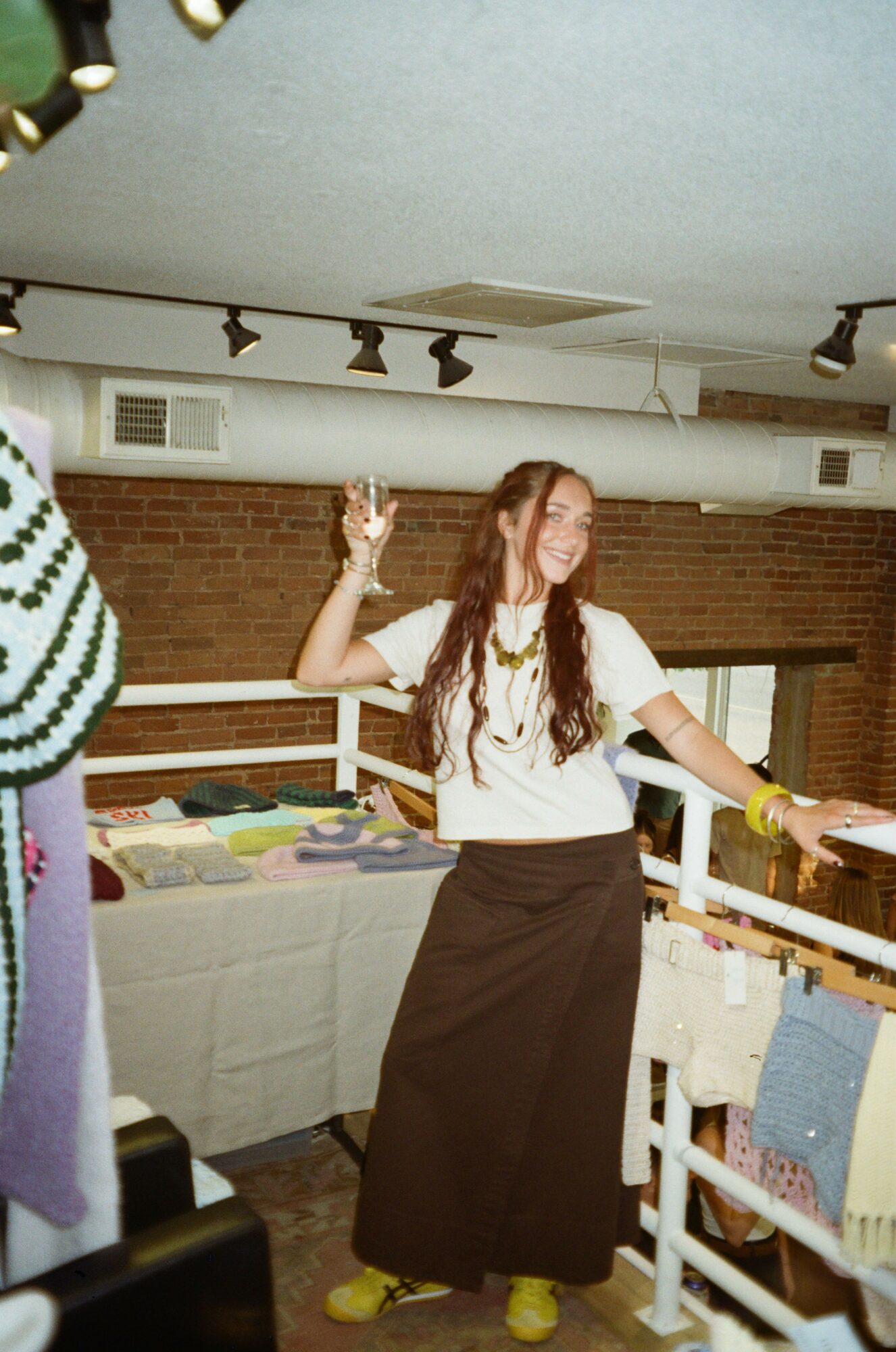 Person with long hair standing in a room, holding a phone, with shelves and a brick wall in the background.
